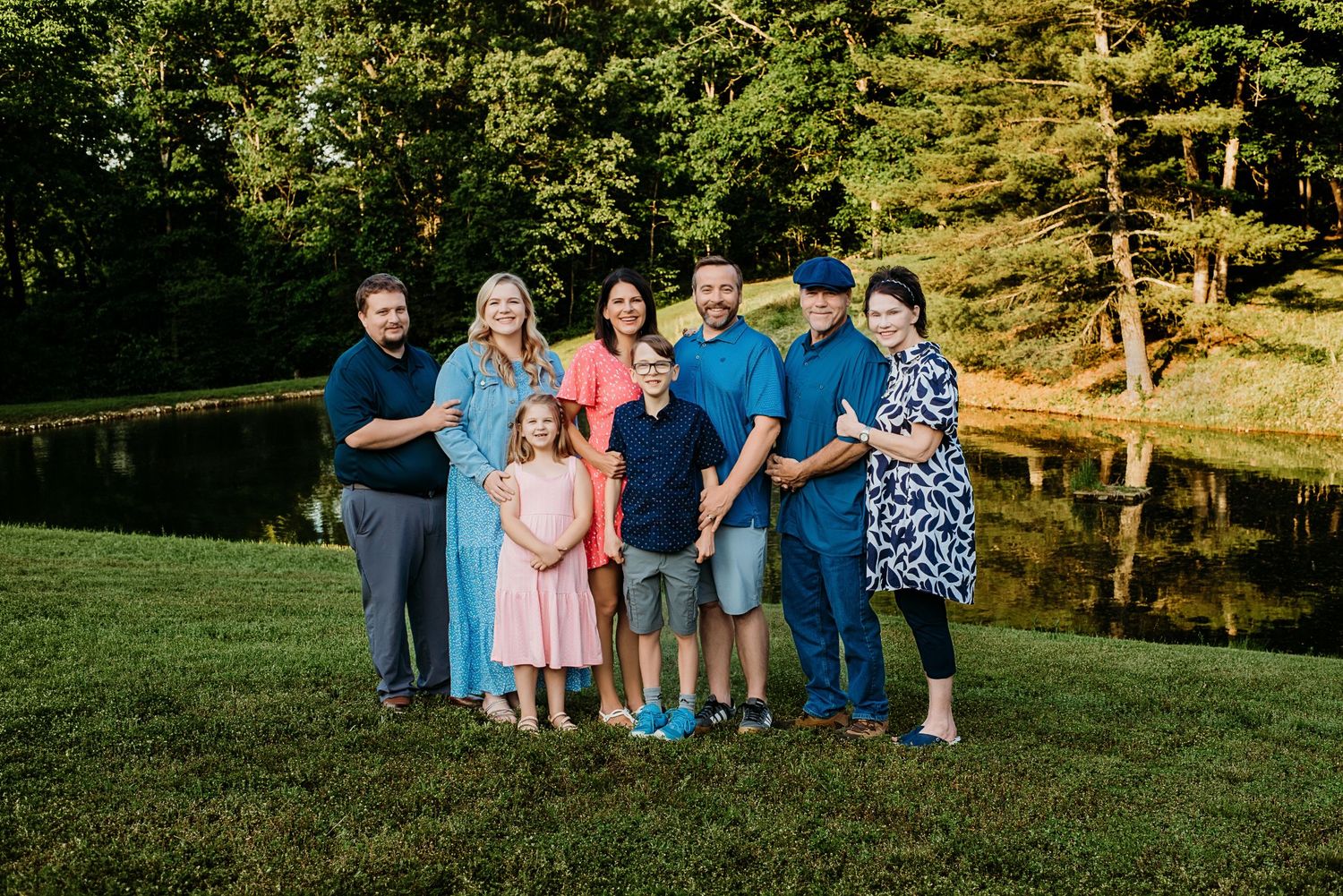 Family of eight smiling outdoors near a pond on green grass during golden hour in a lush park setting.