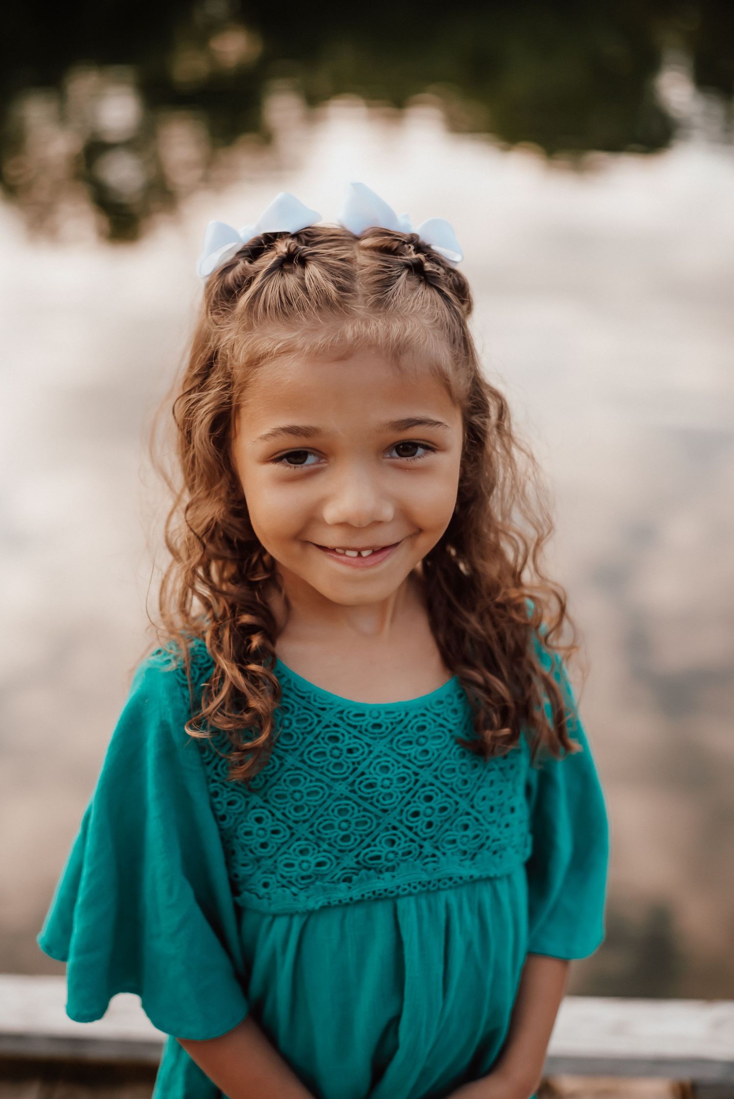 Young girl with curly hair and blue bow smiling, wearing a teal dress outdoors.