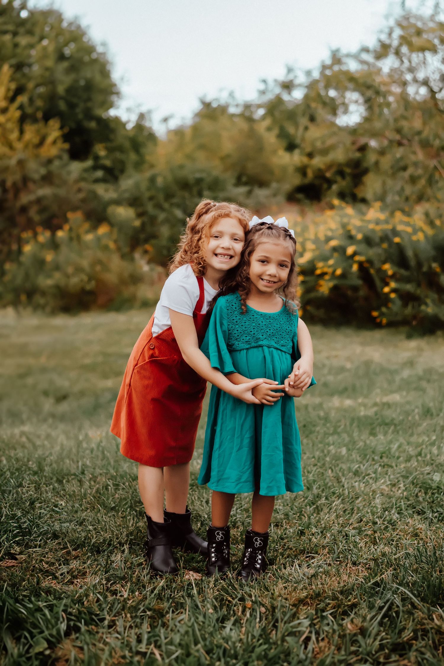 Two young girls hugging and smiling outdoors in a green park, wearing red and teal dresses with black boots.