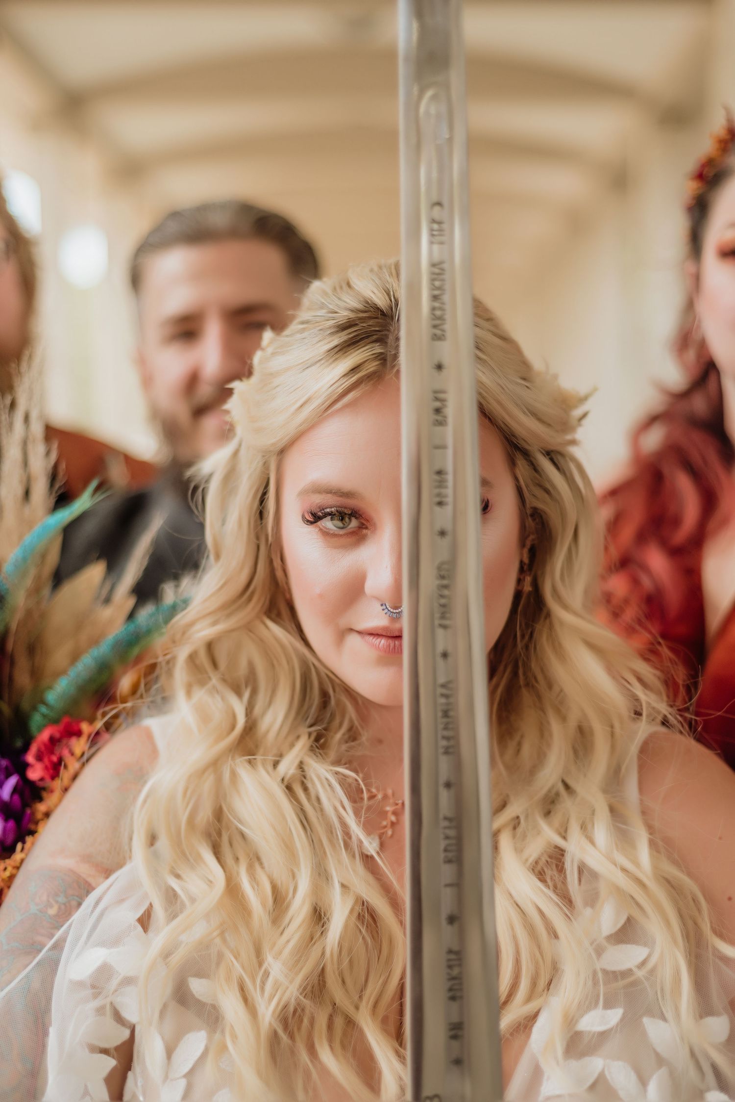 Blonde bride with nose ring peers behind a unity candle, with wedding party members visible in the background.