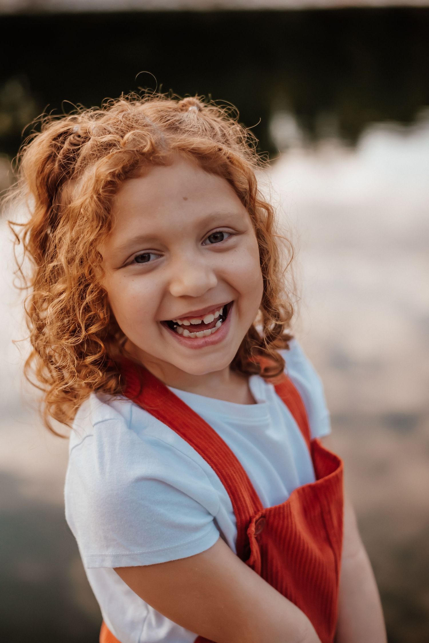 A cheerful young girl with curly red hair smiles brightly, wearing red overalls and a white shirt outdoors.