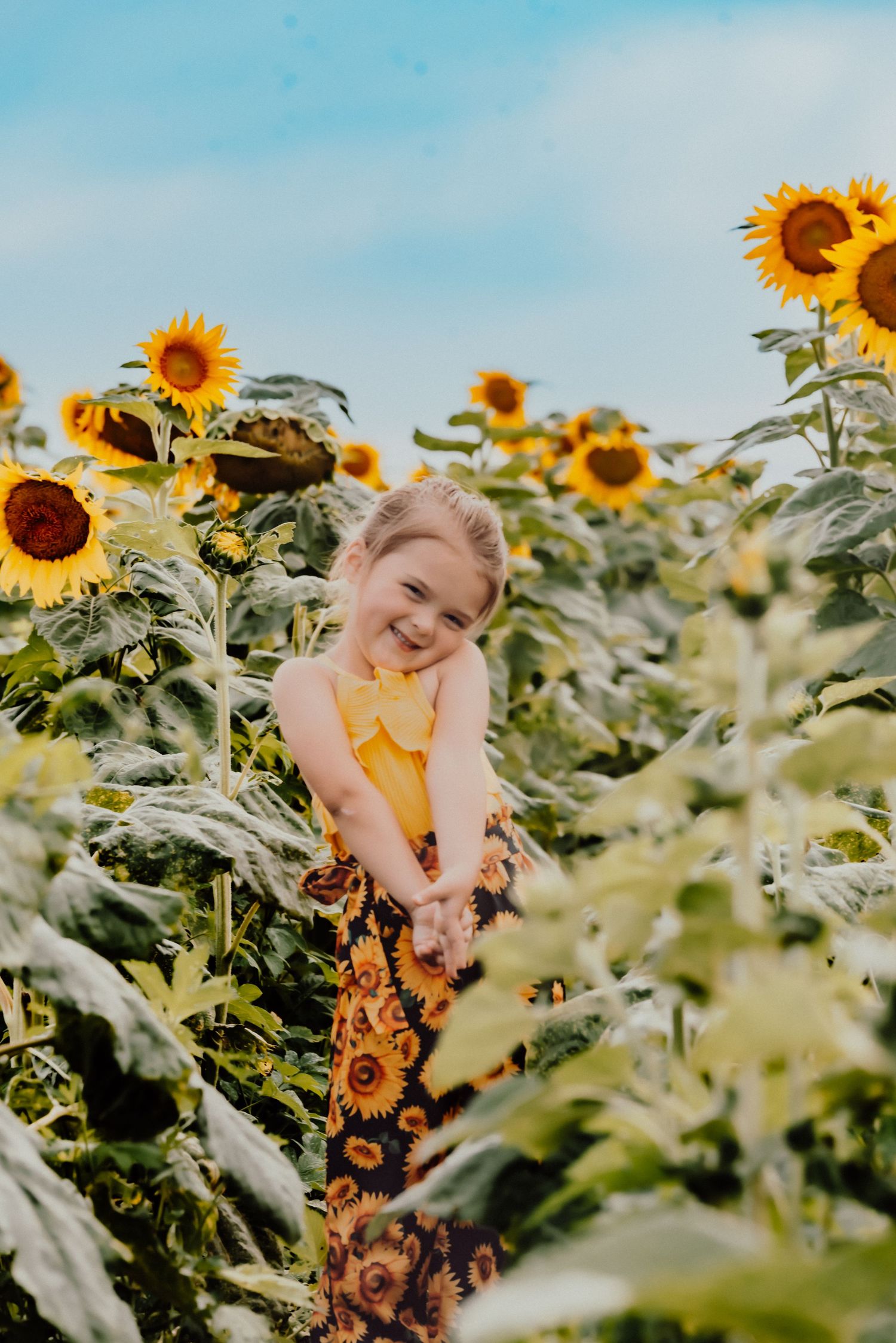 Child in yellow dress standing among tall blooming sunflowers.