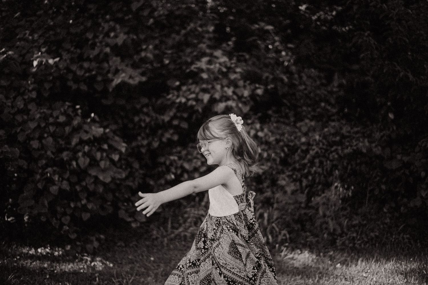 Black and white photo of a young girl in a patterned dress twirling outdoors with trees in the background.