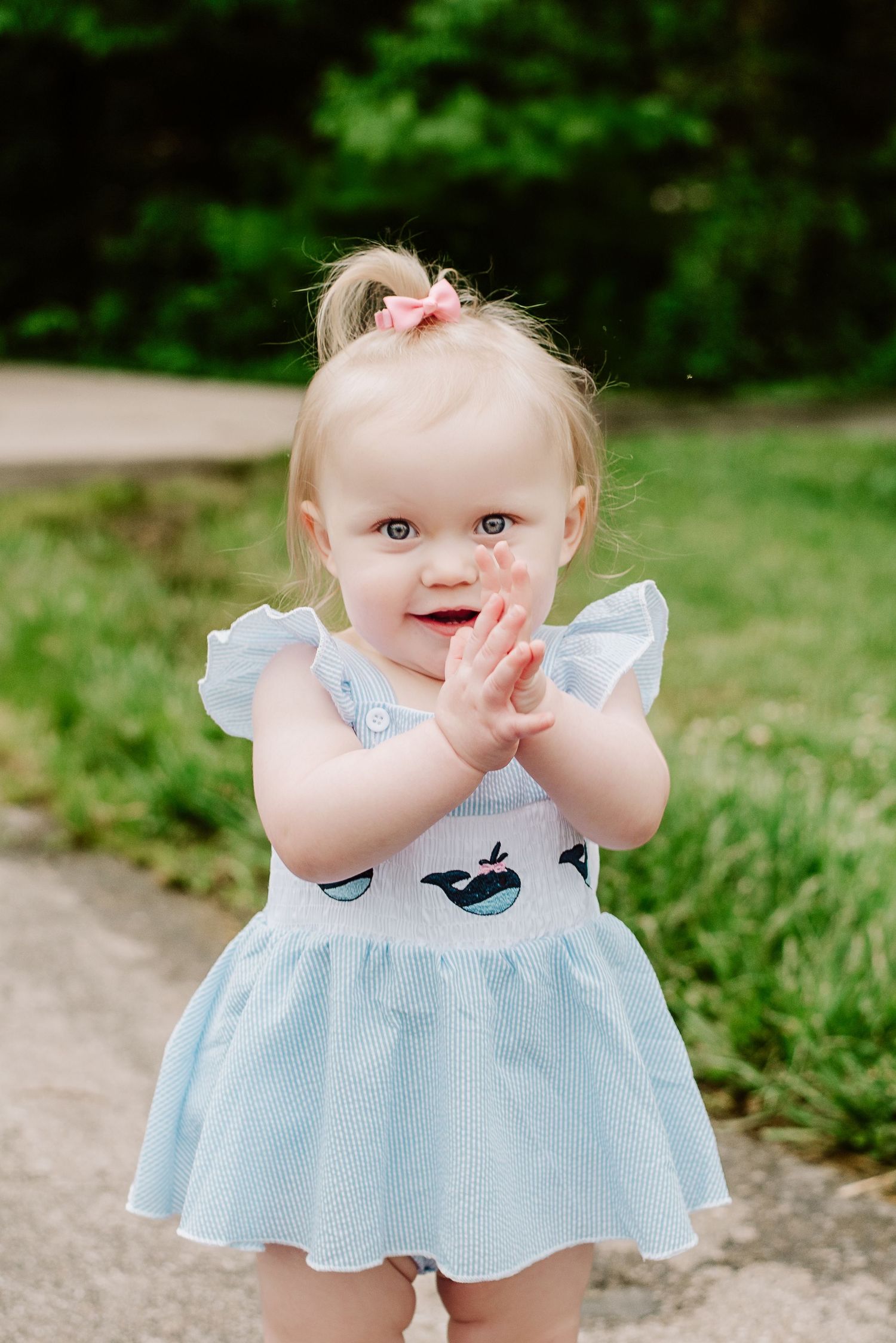 Adorable toddler girl in light blue whale dress with pink bow clapping hands outdoors in green park setting.