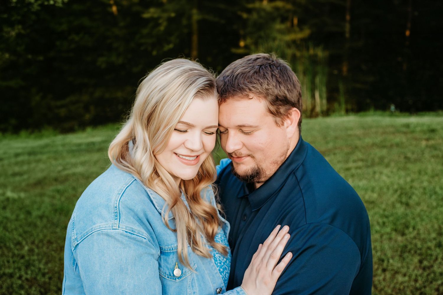 Couple sharing a tender moment outdoors, woman in denim jacket smiling with heads together in a green field.