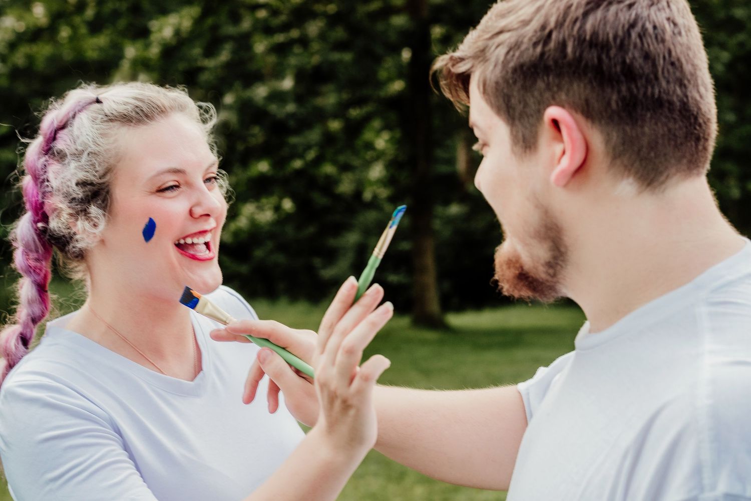 A laughing couple painting each other's faces with blue paint in a garden.