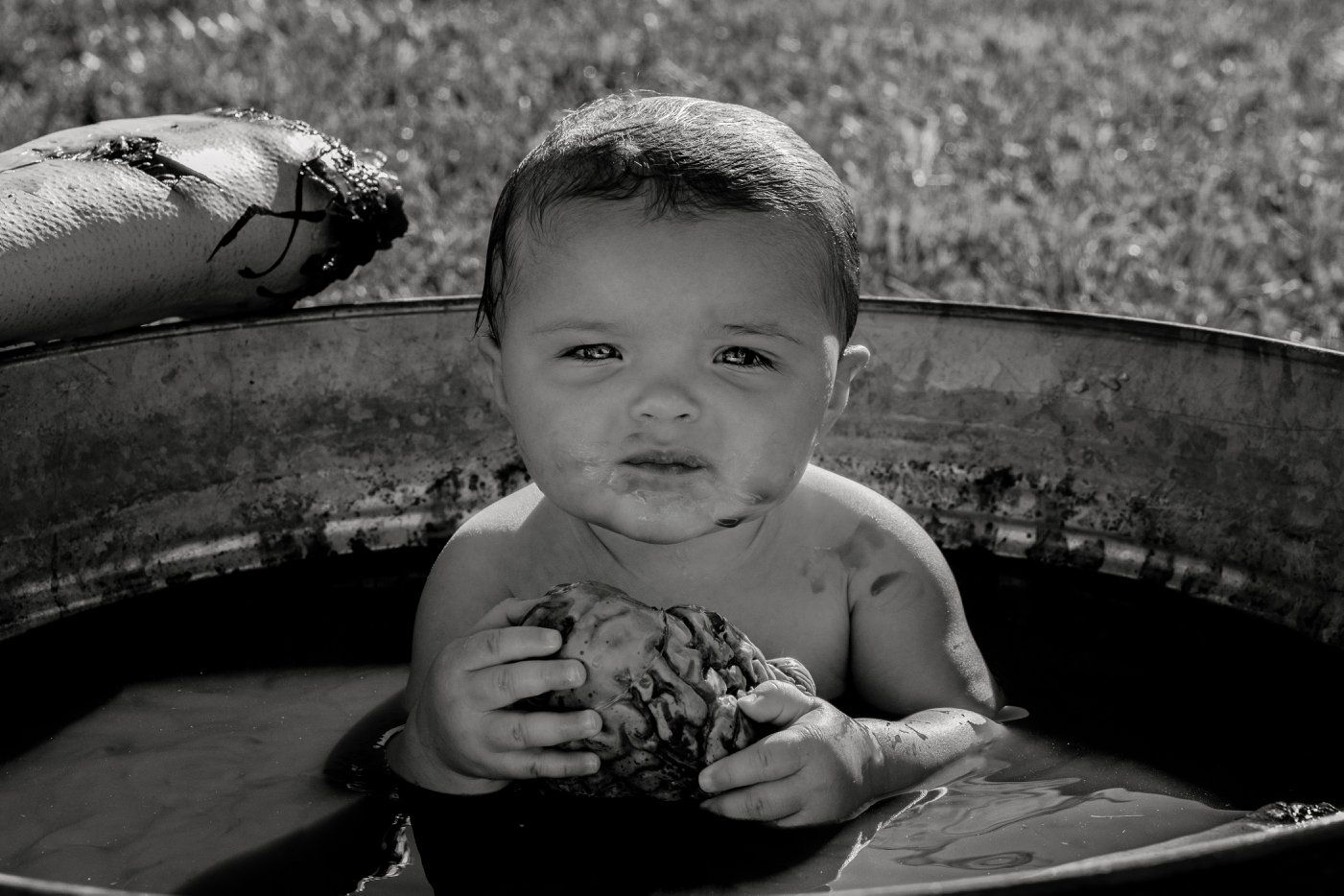 A baby sits in a metal tub holding an object in black and white photography.