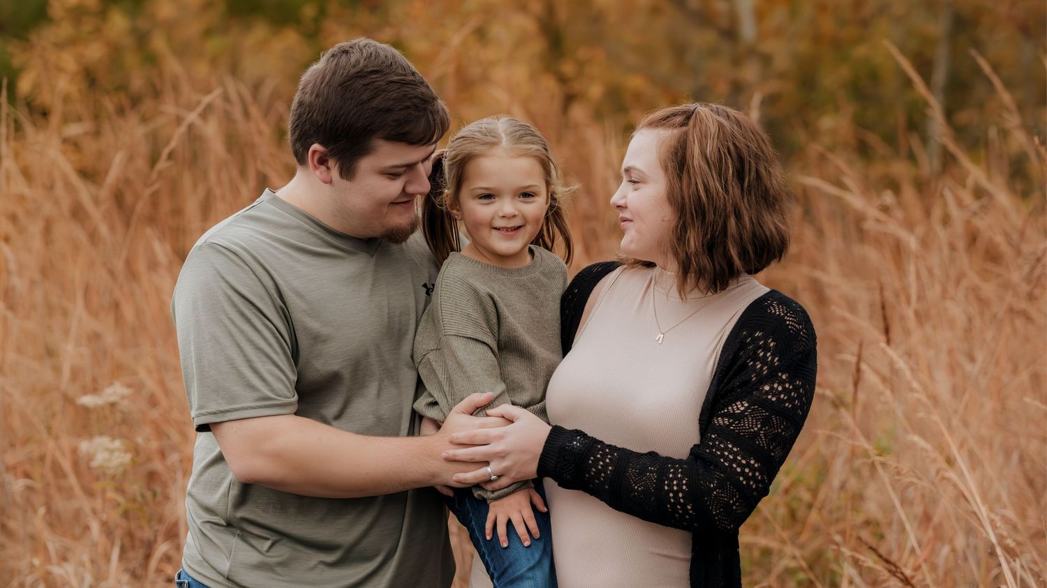 A happy family of three posing outdoors in a golden autumn field, parents smiling at their young daughter.
