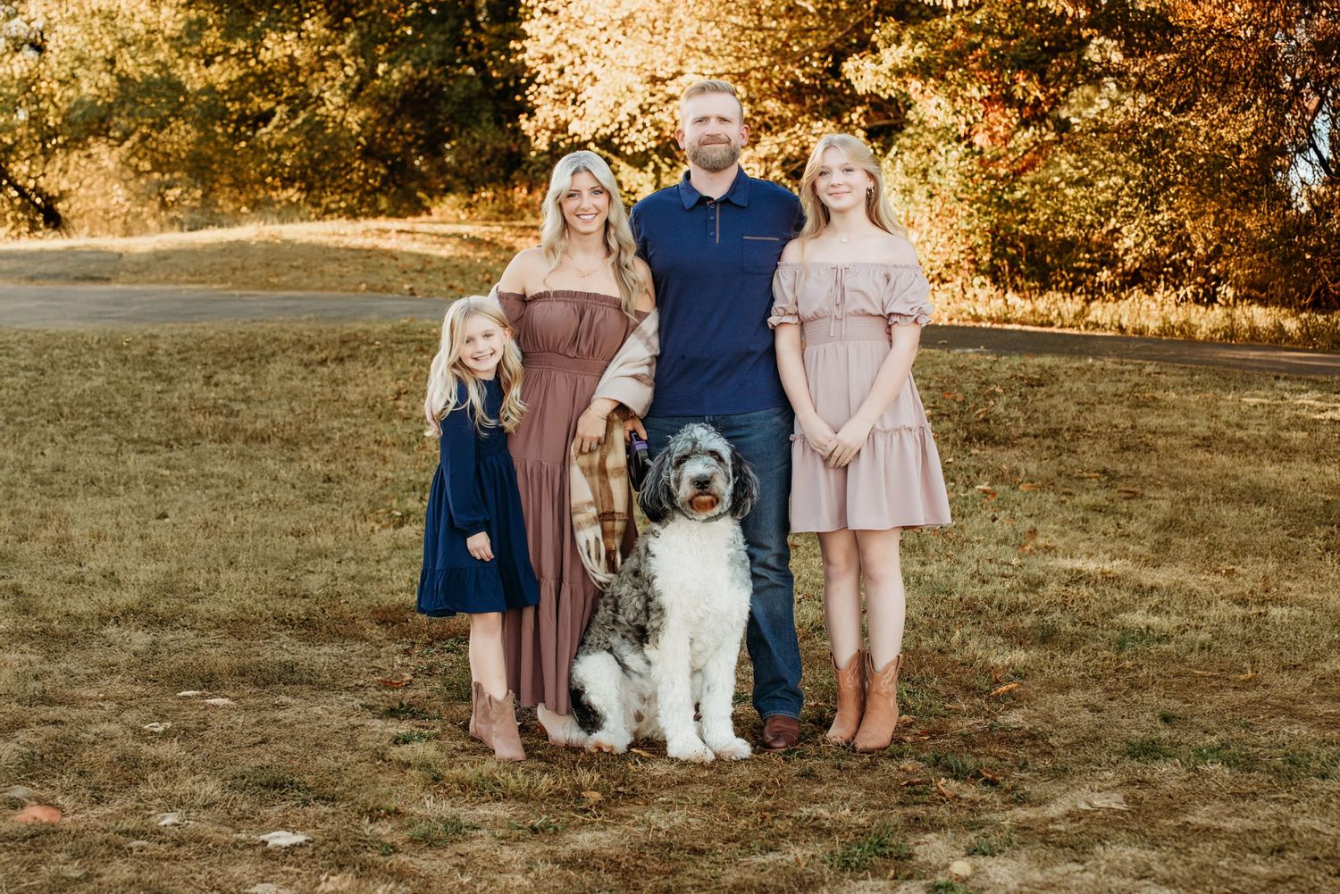 Family of four with a fluffy dog posing outdoors in autumn park with golden foliage backdrop.