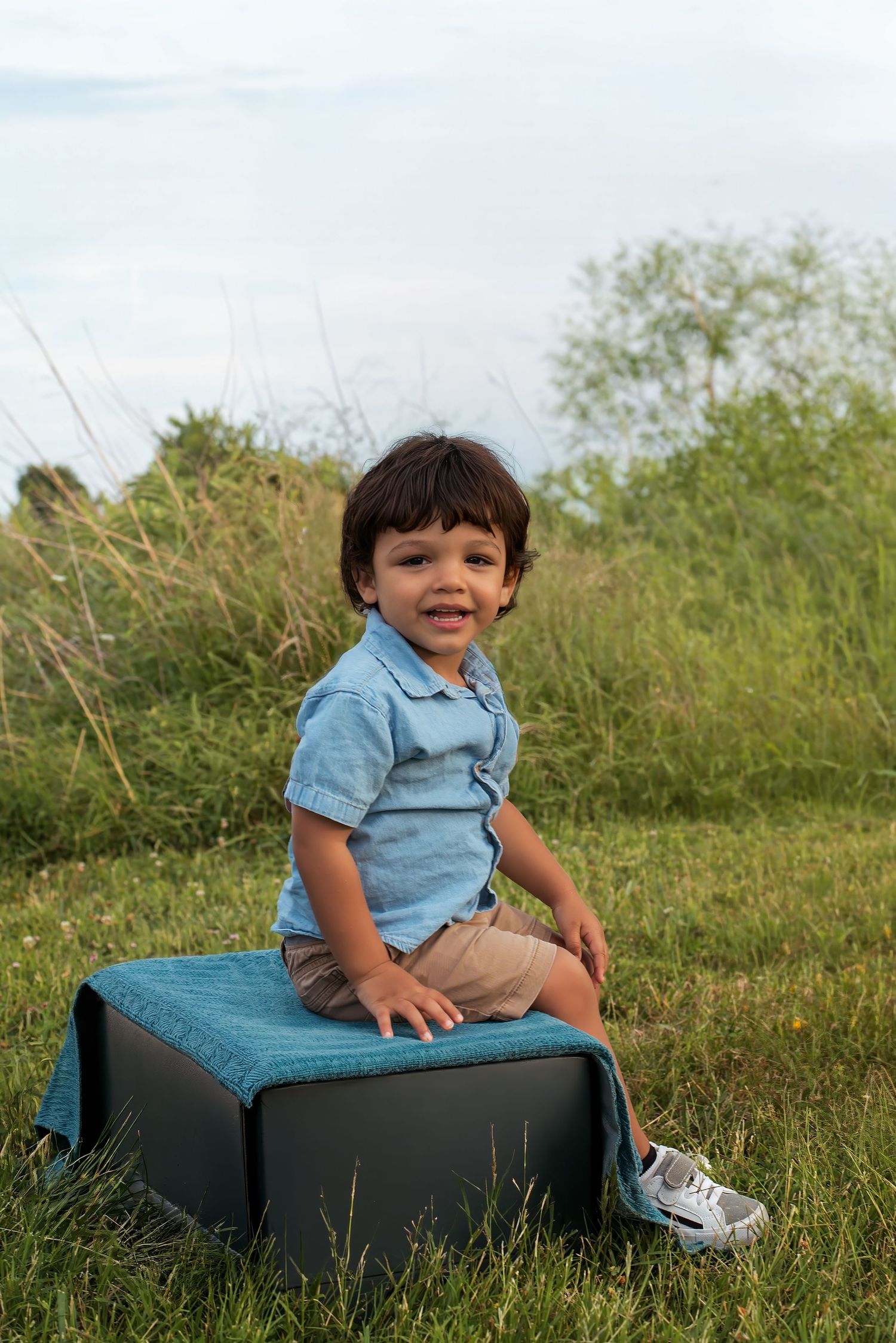 A smiling toddler boy in a blue polo shirt sits on a teal ottoman in a lush green outdoor field.
