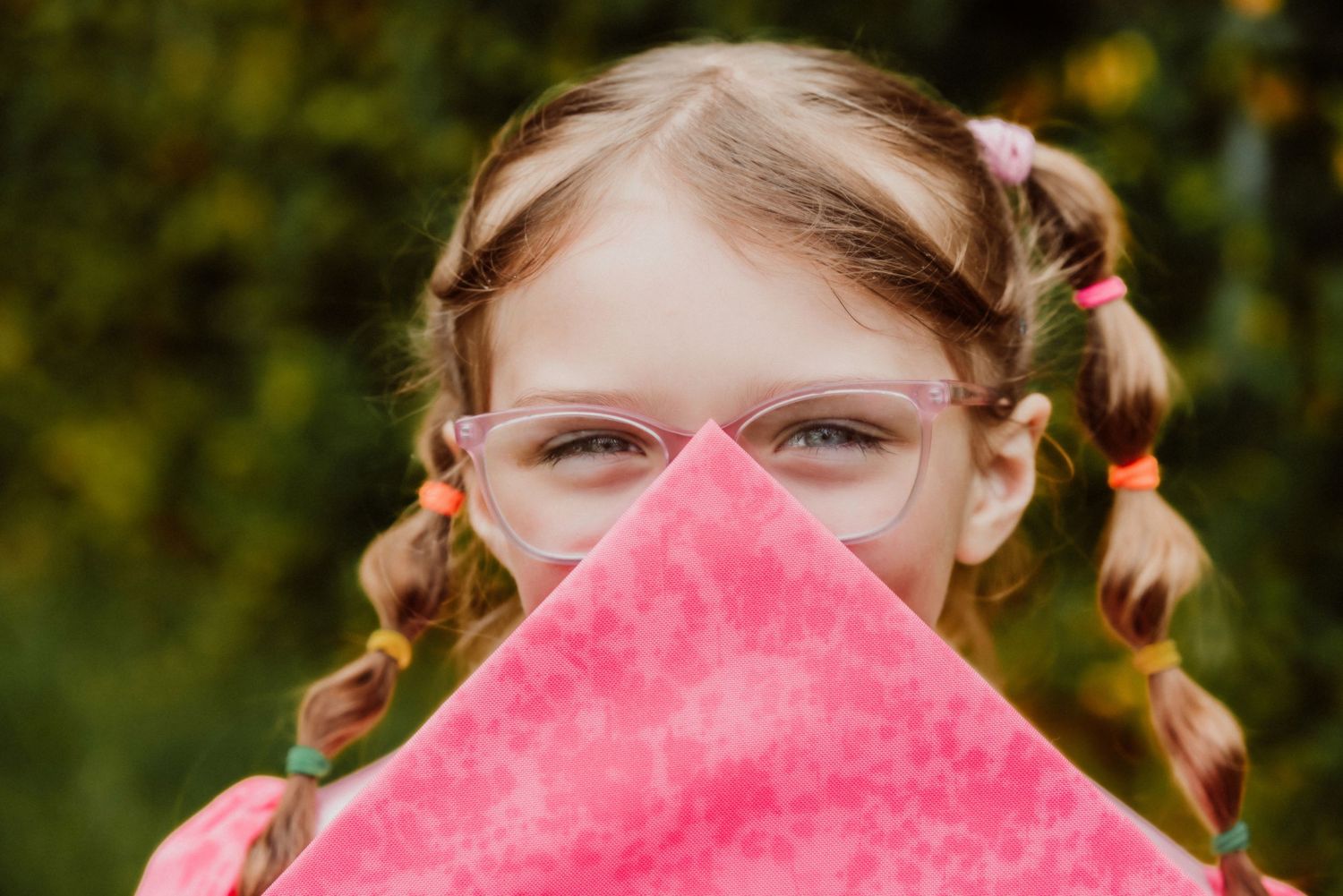A playful portrait showing braided pigtails and glasses peeking over pink fabric.