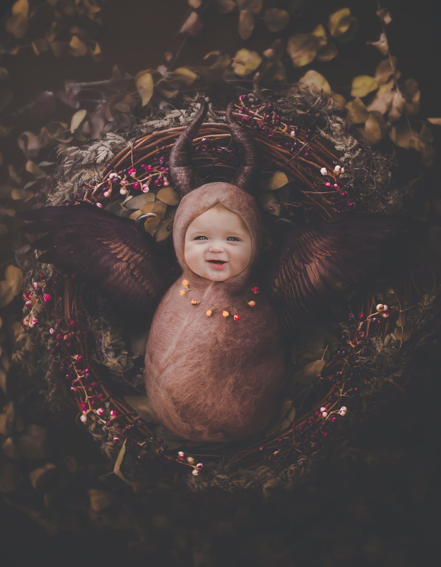 Smiling newborn baby wrapped in brown swaddle with deer antler bonnet, nestled in autumn wreath with red berries.
