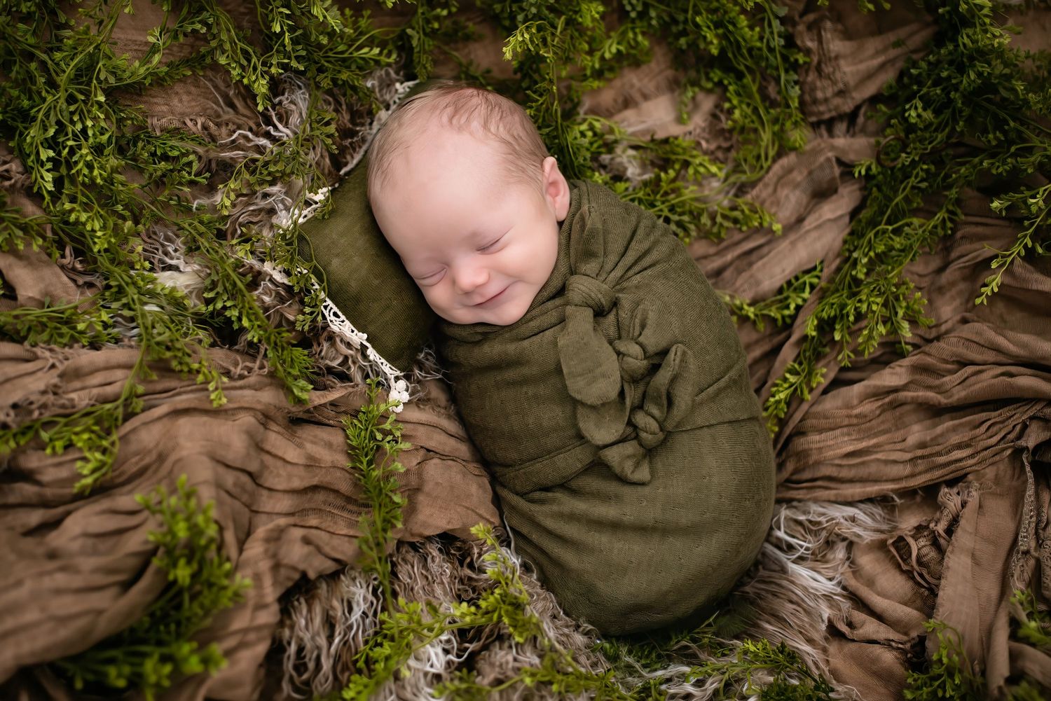 Smiling newborn baby wrapped in green swaddle cloth, nestled among moss and brown textured fabric in nature-inspired setting.