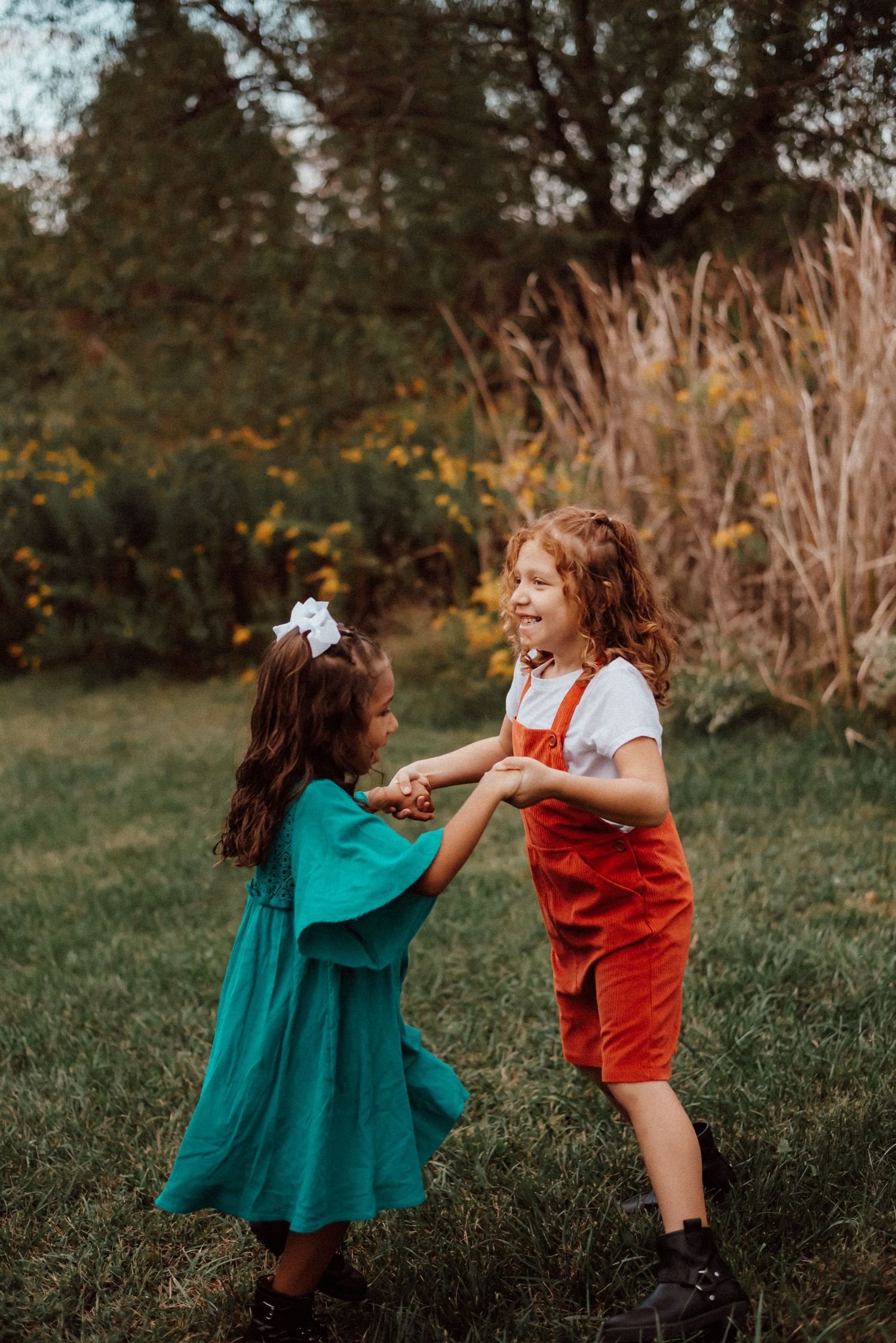 Two young girls in colorful outfits hold hands and dance together on a green lawn with wildflowers in the background.
