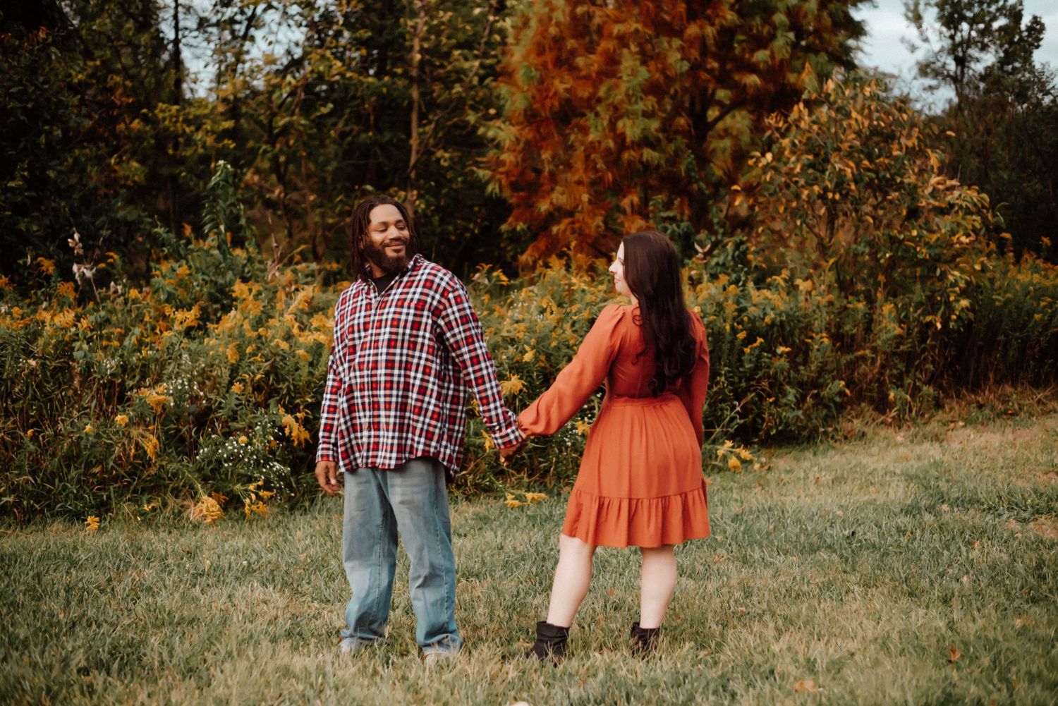 A couple holds hands while walking through a vibrant autumn landscape, smiling at each other in coordinating outfits.