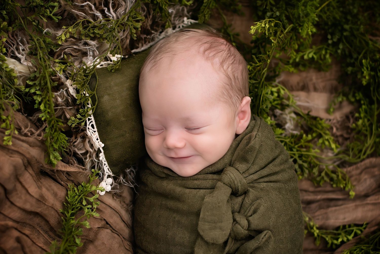 Smiling newborn baby wrapped in green swaddle nestled among moss and wood in a nature-themed photo.