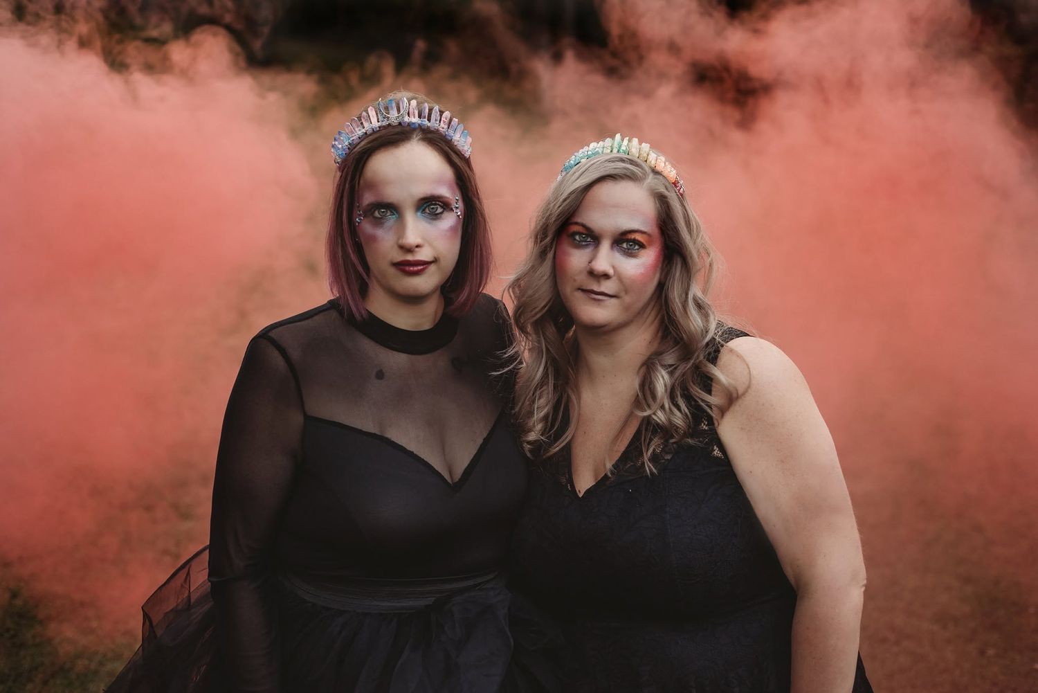 Two women in black dresses and tiaras pose against a dramatic pink smoke backdrop.