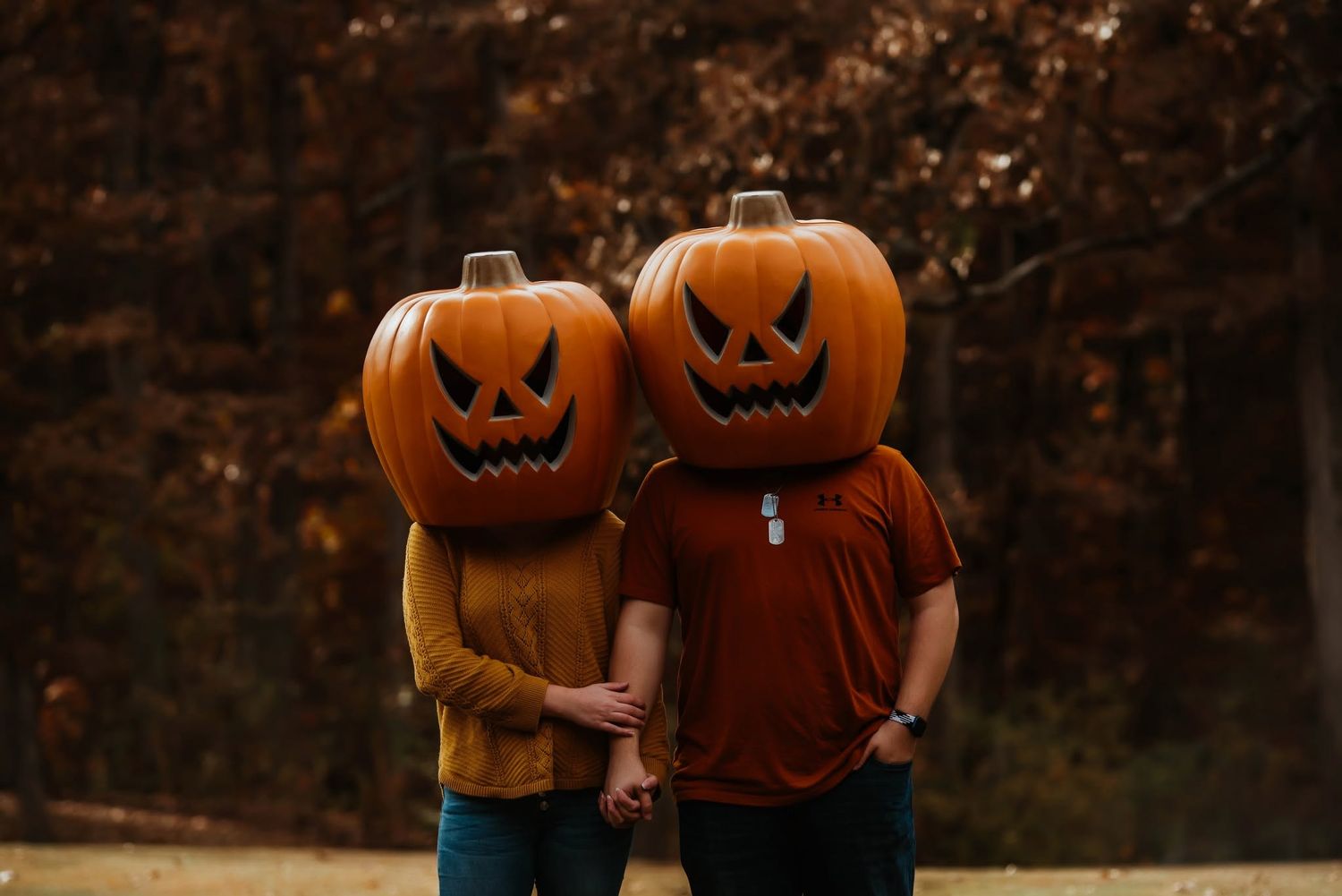 Couple wearing large carved jack-o-lantern pumpkins on their heads holding hands outdoors during autumn fall season.