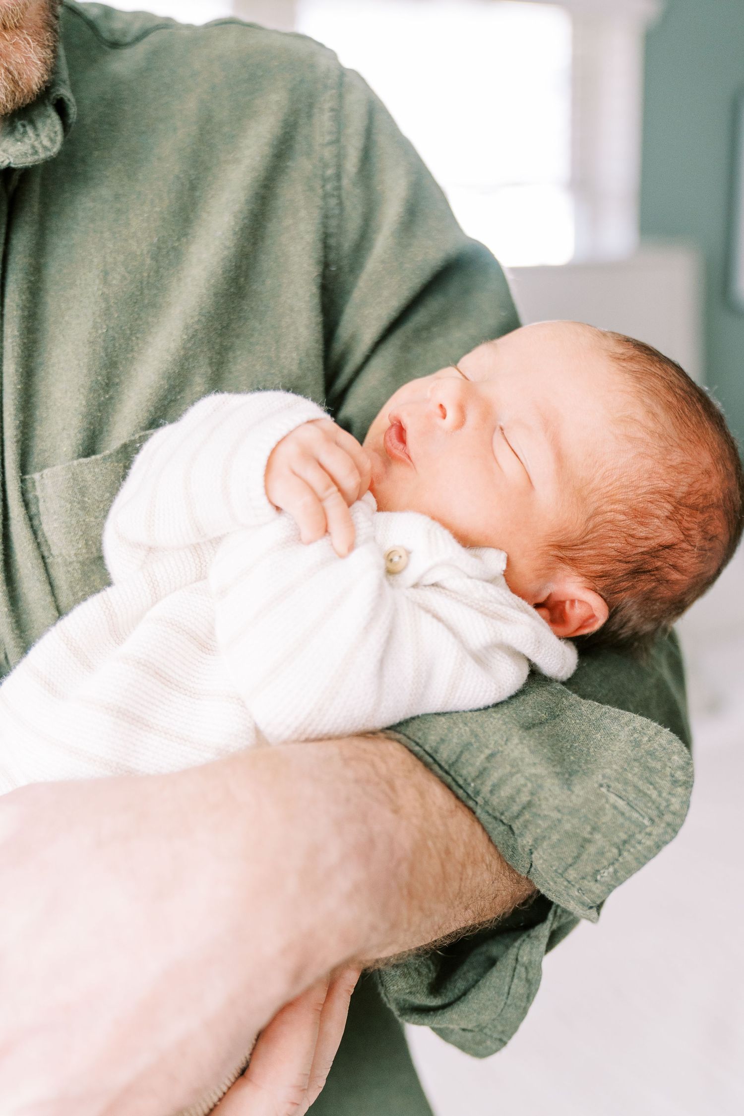 A peaceful sleeping newborn baby wearing white clothing rests in someone's arms wearing a sage green sweater.