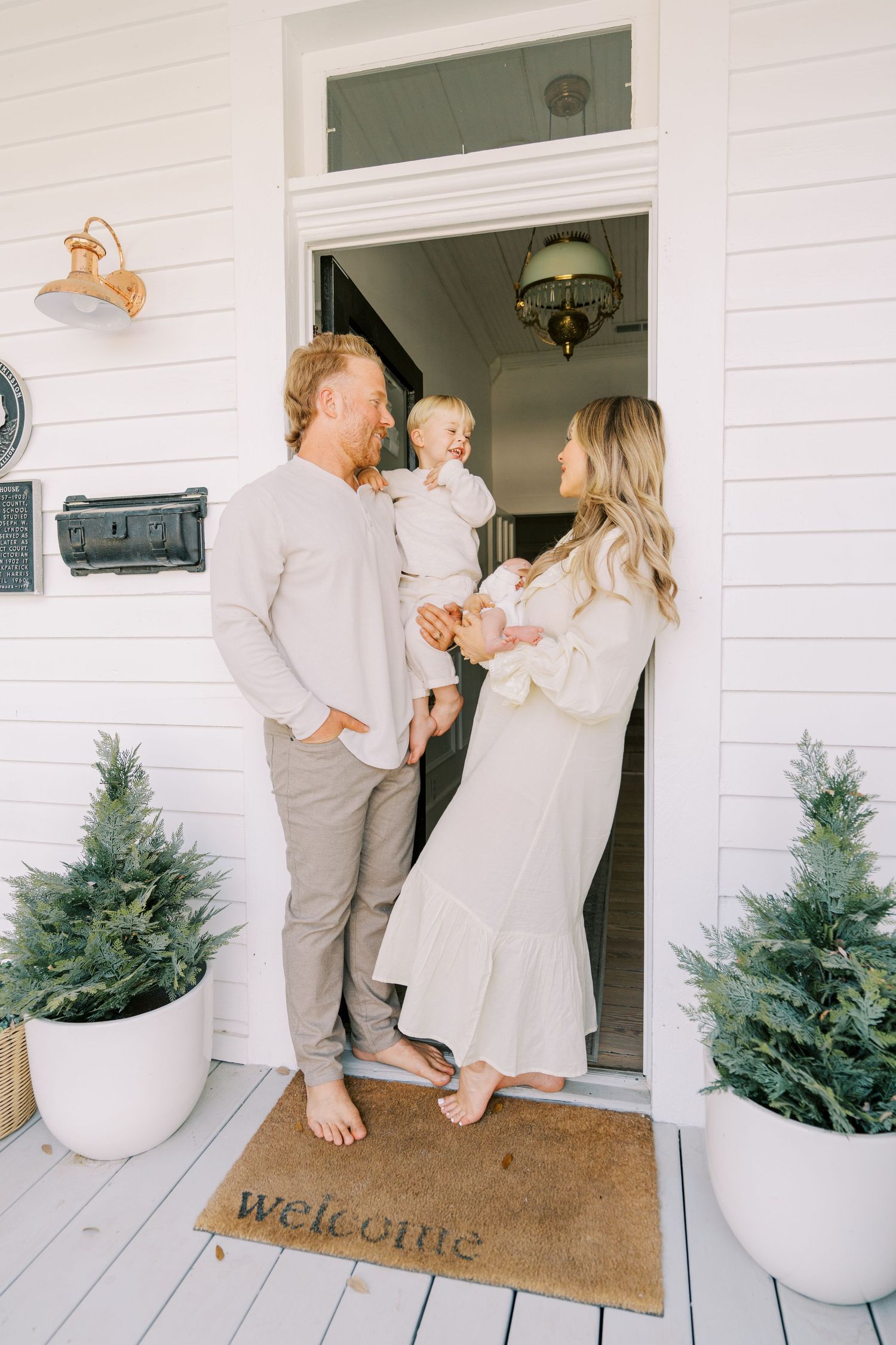 Family in neutral tones stands at front door with welcome mat and potted evergreens.