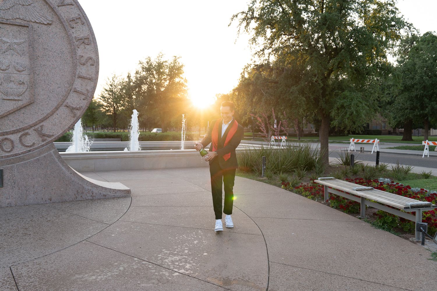 Silhouette of a person walking through a park at sunset with fountains and benches in the background.