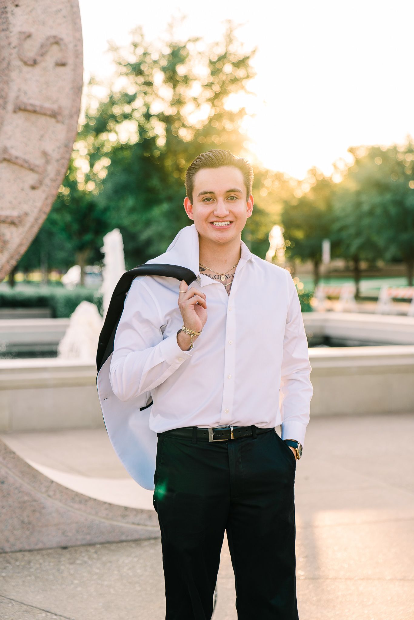 A person in formal attire stands outdoors at sunset wearing a white dress shirt and black pants.