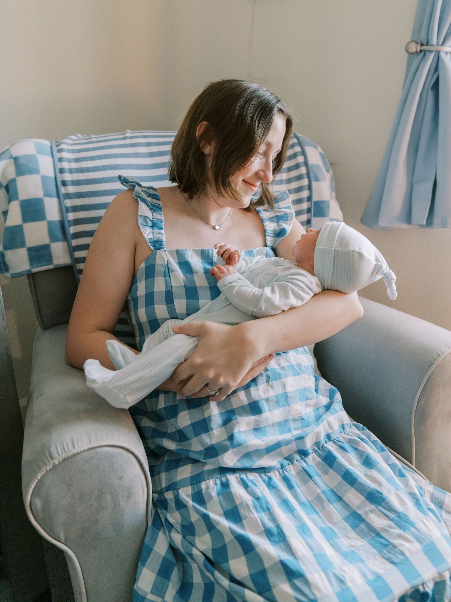 Parent in blue gingham dress tenderly holding and feeding newborn baby in a cozy armchair.