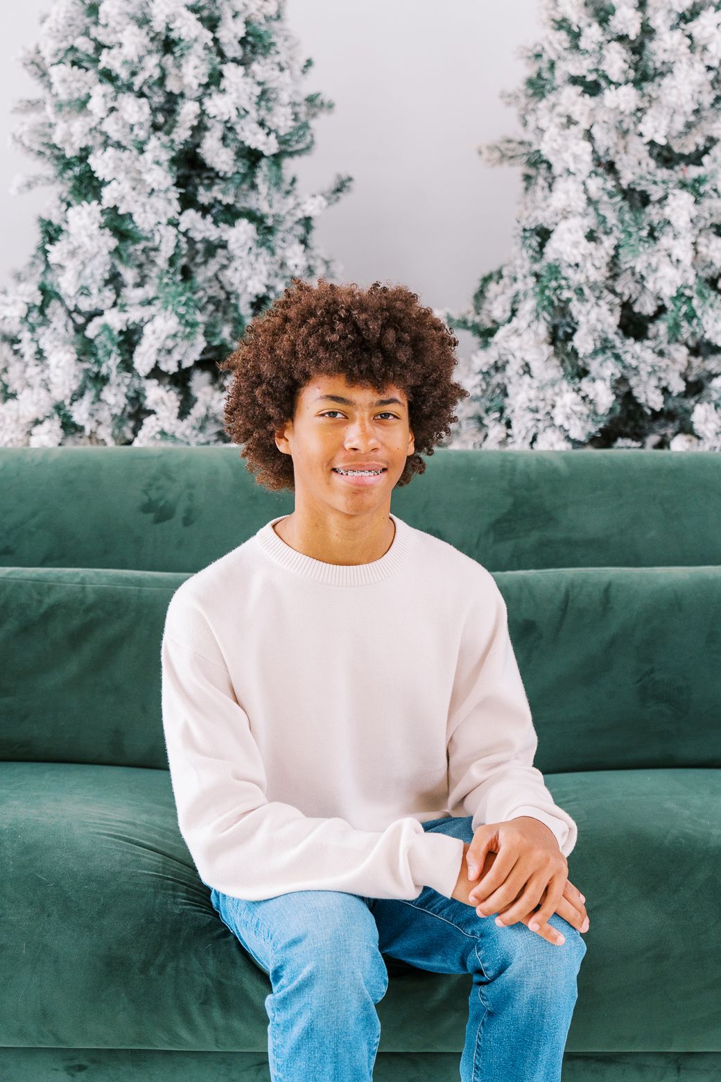 Individual in casual white sweater and jeans seated on emerald couch with snowy Christmas trees as backdrop.