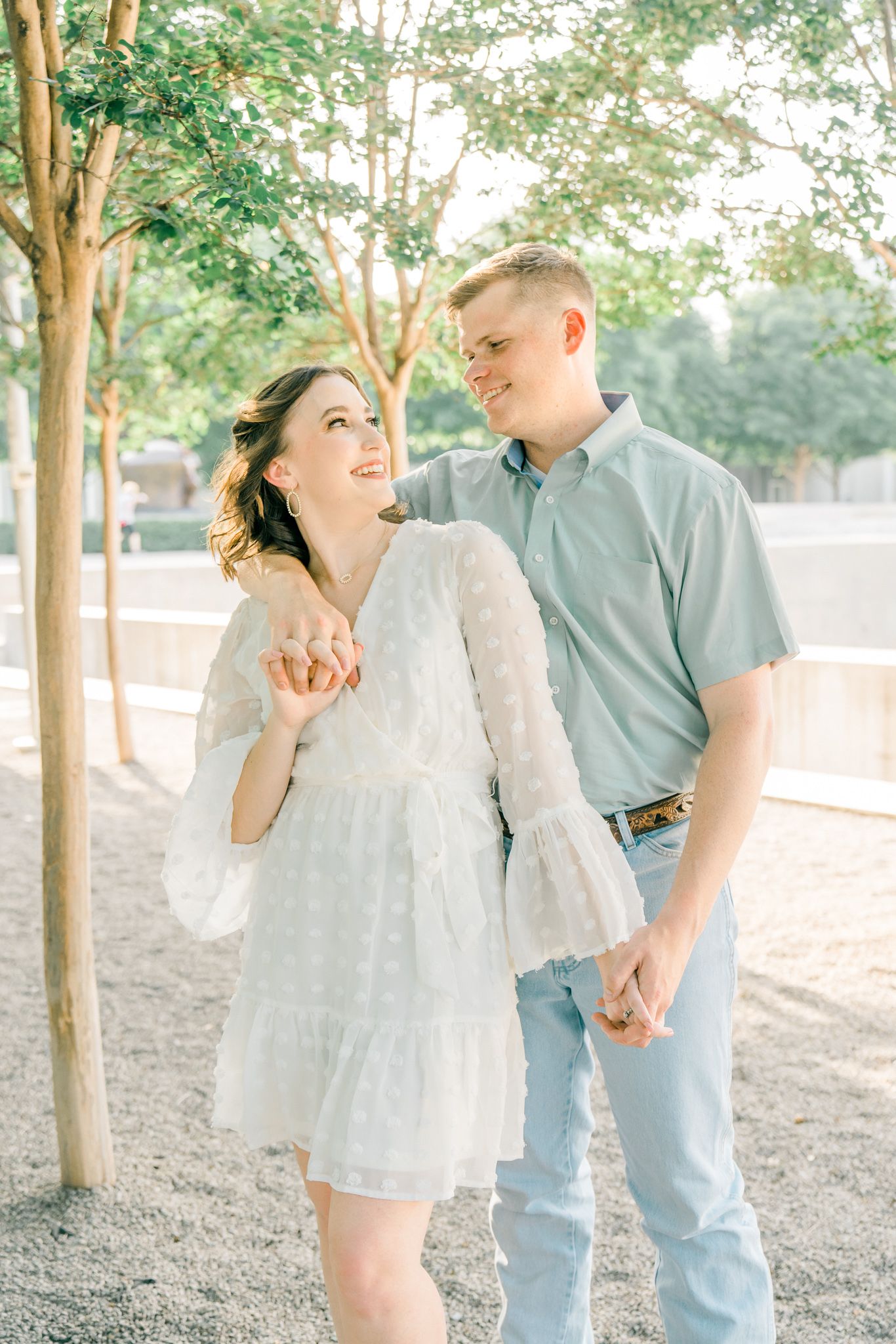 Couple in light colored outfits sharing romantic moments under a canopy of trees.