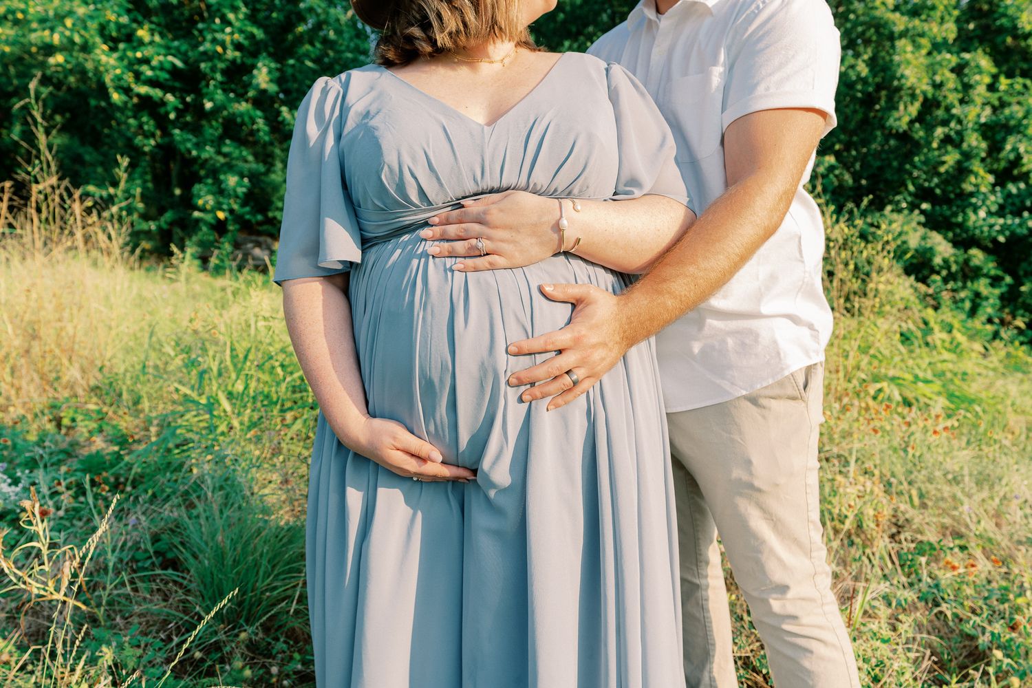 A couple in soft gray and beige attire embraces during an outdoor photoshoot in a lush natural setting.