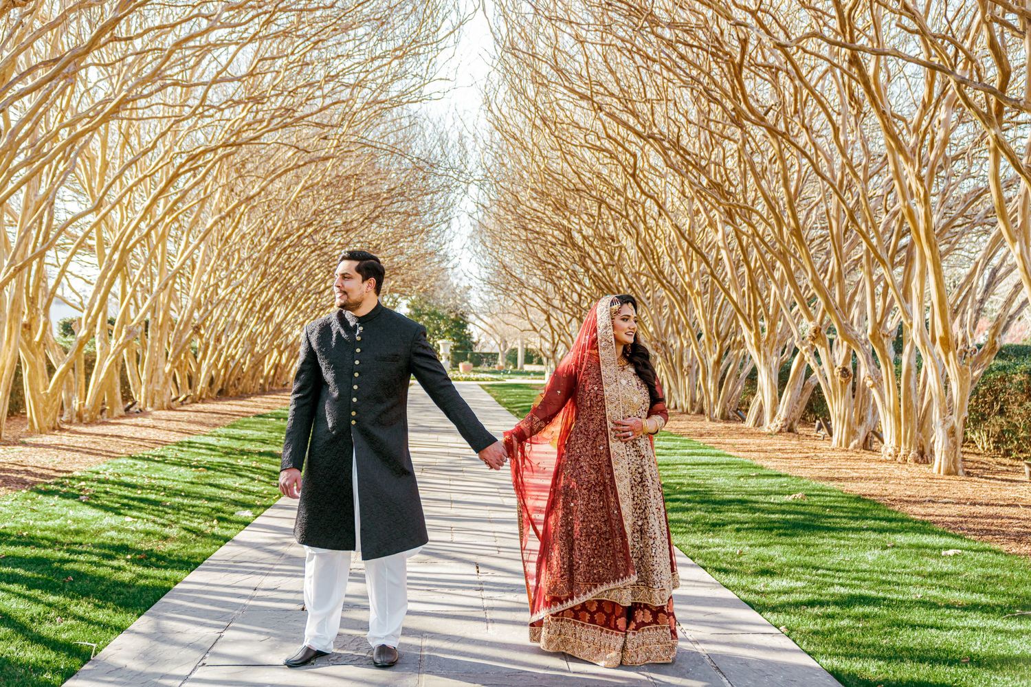 A couple walks hand in hand through a tree-lined path during their wedding photoshoot.