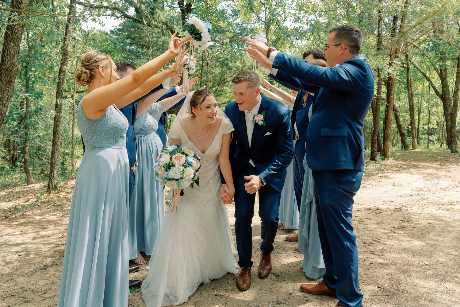 Wedding party in light blue dresses and navy suits celebrating with a champagne toast in a wooded outdoor setting.