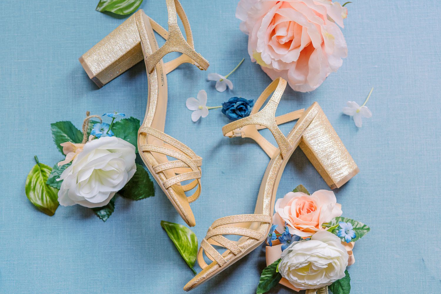 Elegant nude strappy wedding heels surrounded by white and peach roses on light blue surface.
