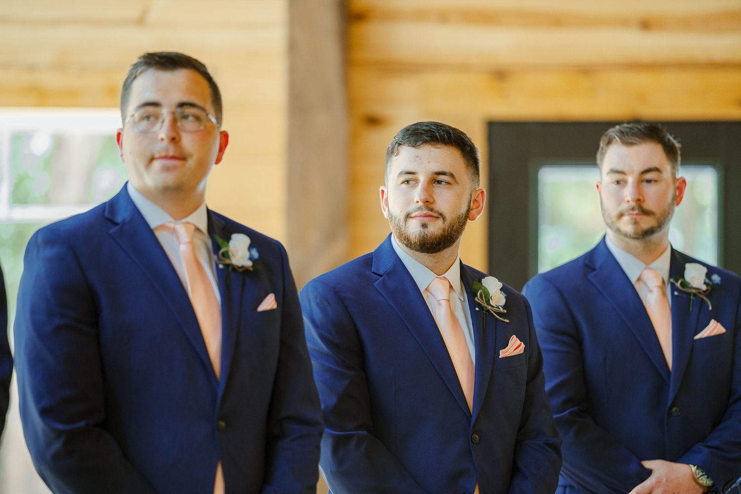 Three groomsmen in matching navy blue suits with pink ties stand together at a wedding ceremony.