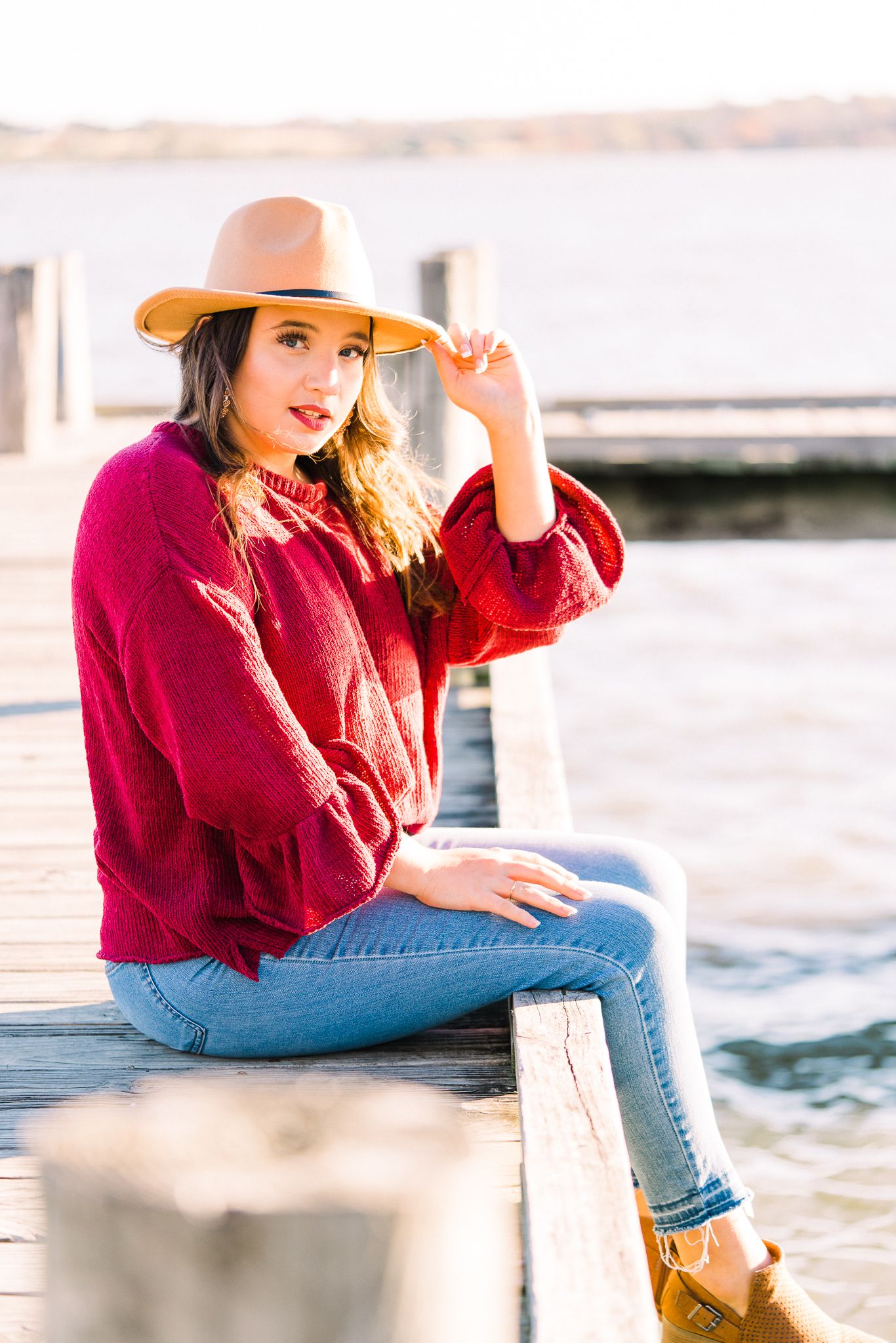 Person in red sweater and straw hat poses on wooden dock with water view at golden hour.