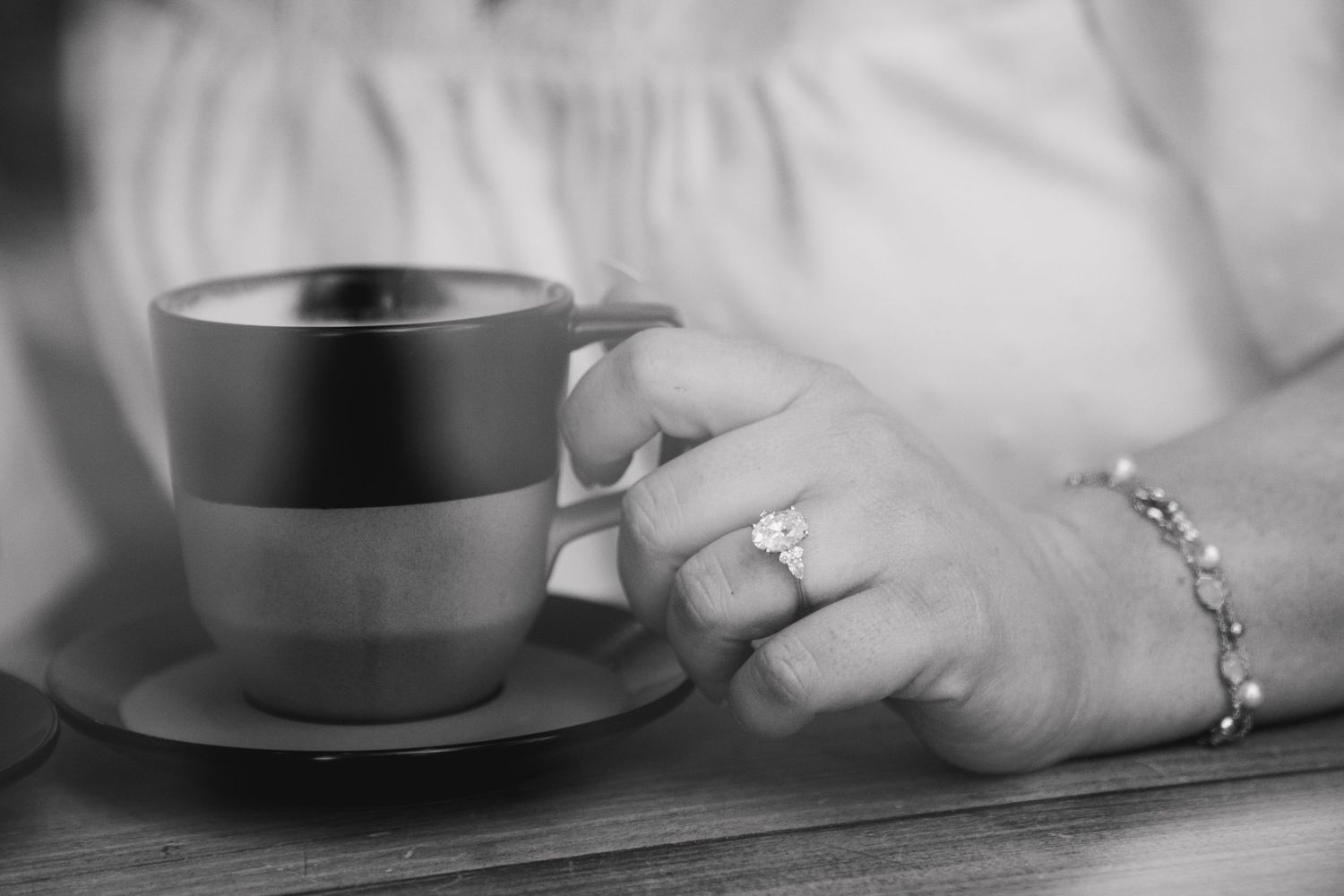 Close-up black and white photo of a woman\u2019s hand holding a coffee cup, wearing a diamond engagement ring.