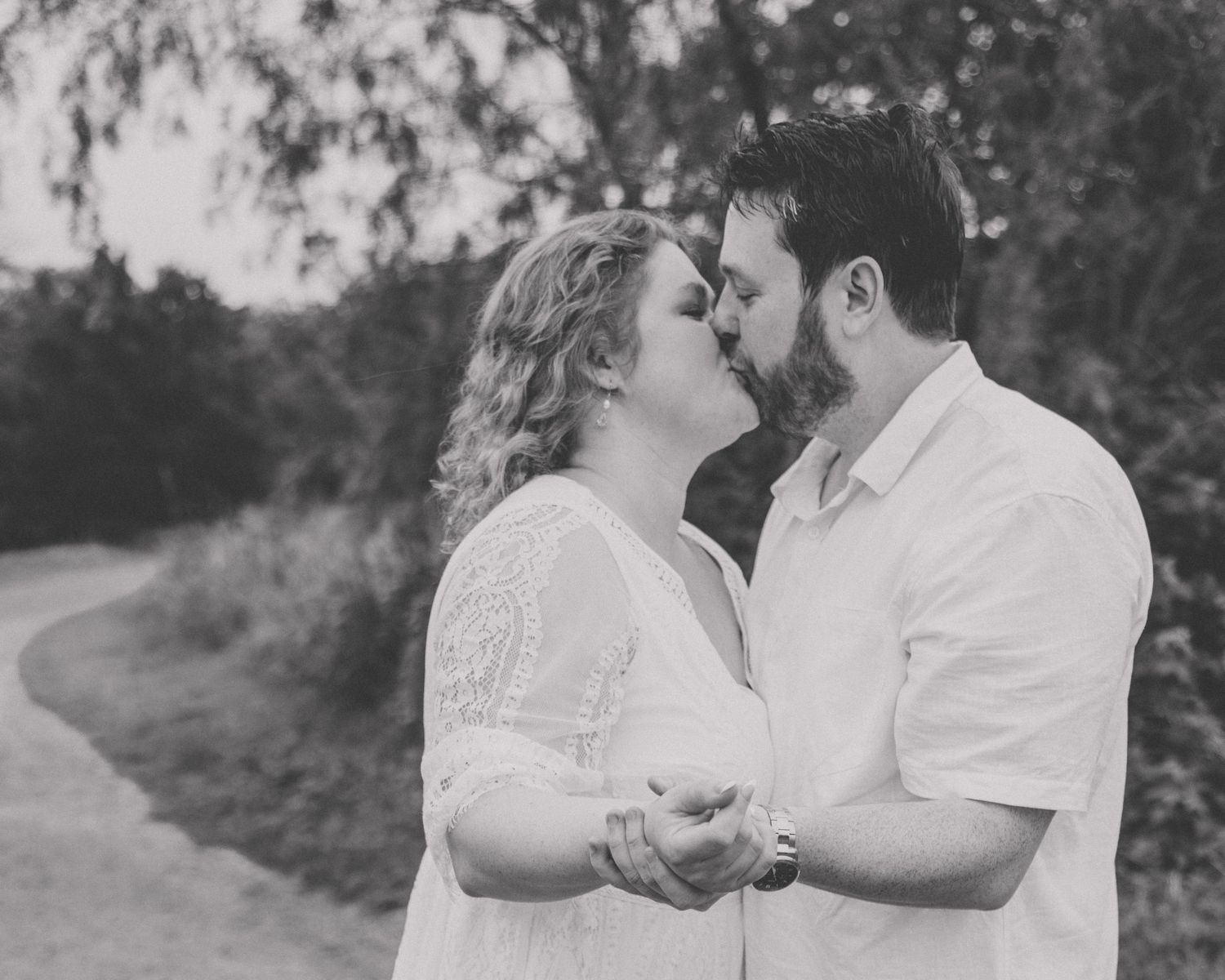 Black and white photo of couple kissing on a scenic outdoor path, holding hands romantically.