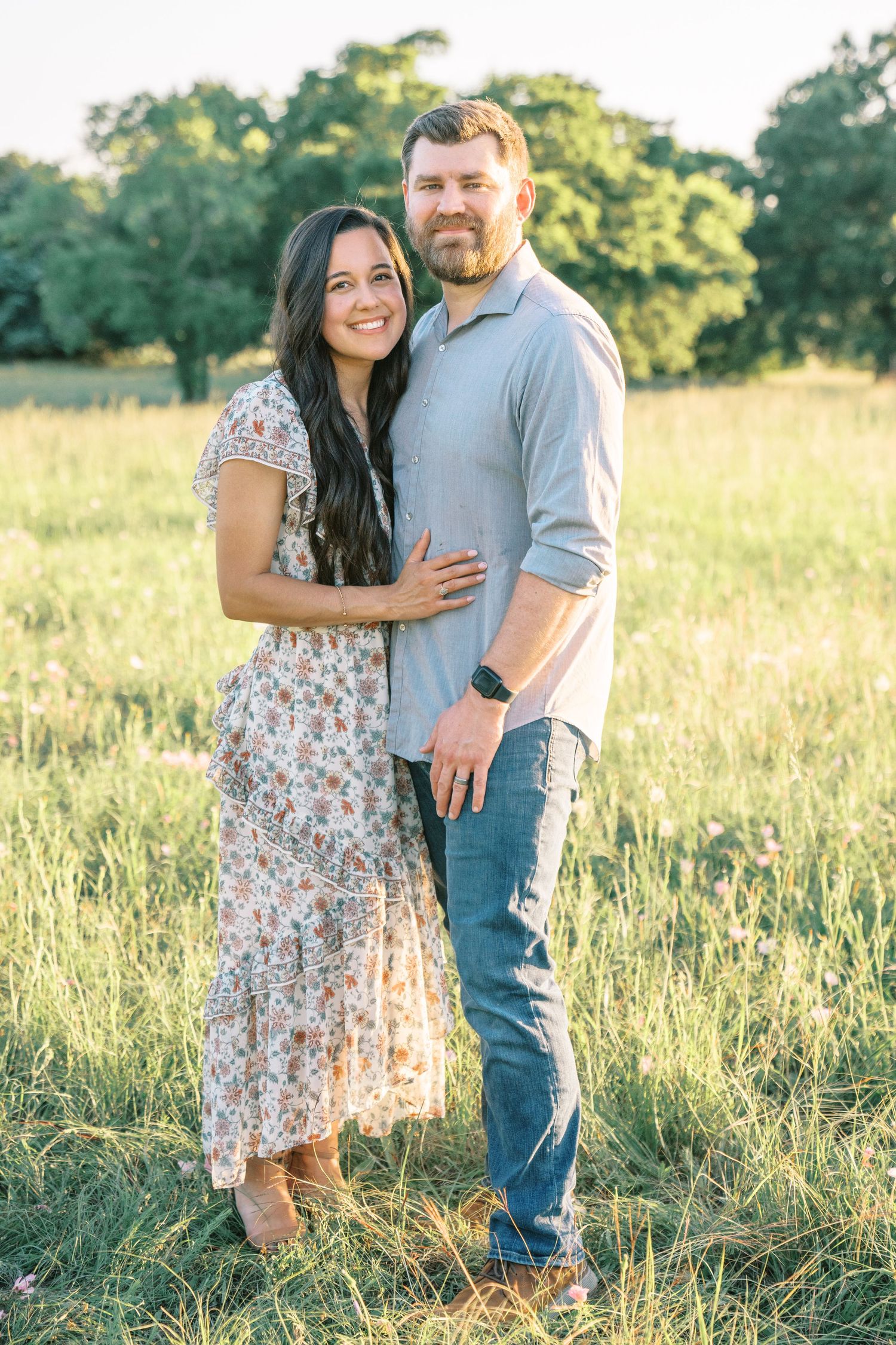A couple embraces smiling in a sunlit green field, woman in floral dress, man in gray shirt and jeans.