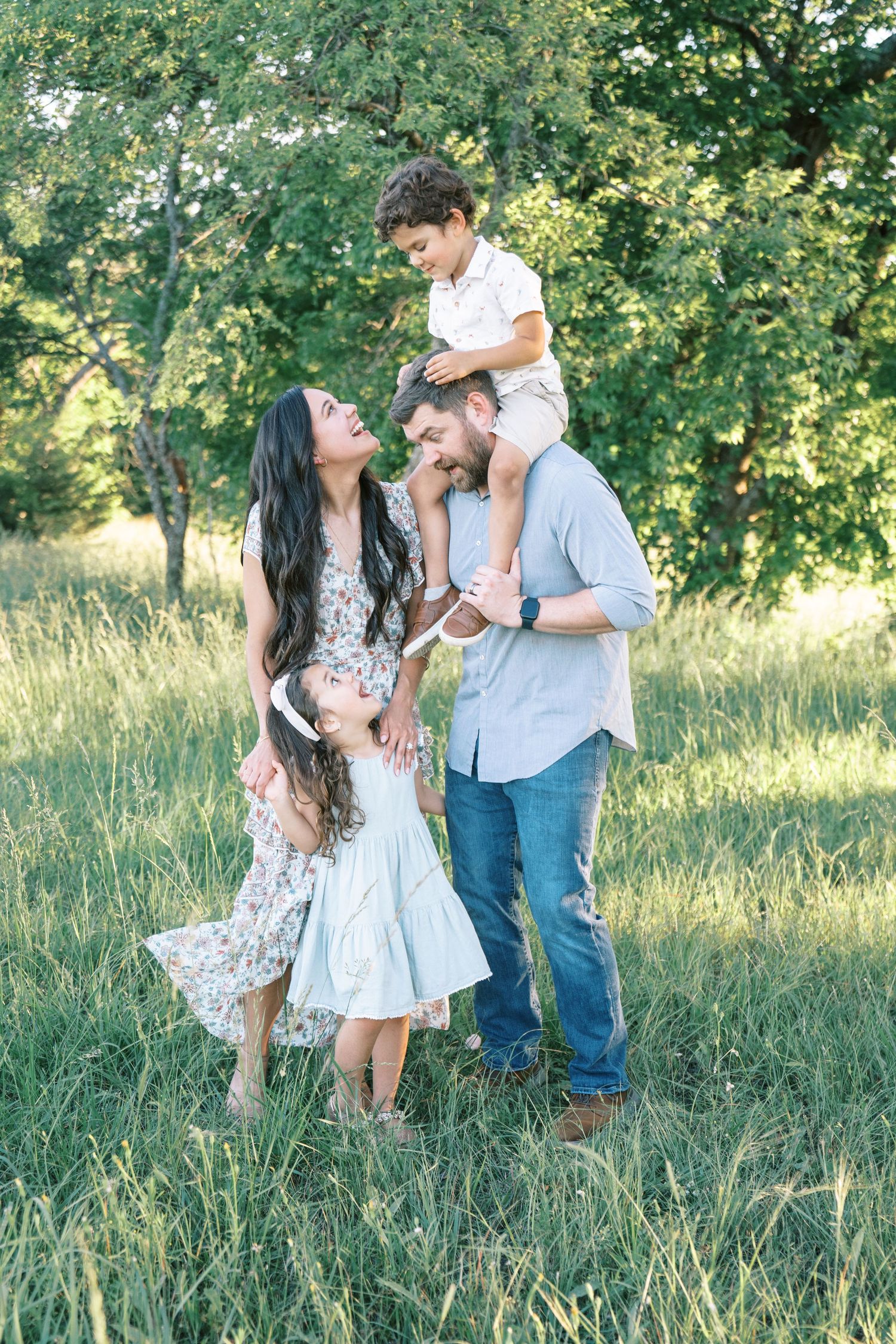 A joyful family of four plays in a green meadow, with a boy on dad's shoulders and a girl twirling nearby.