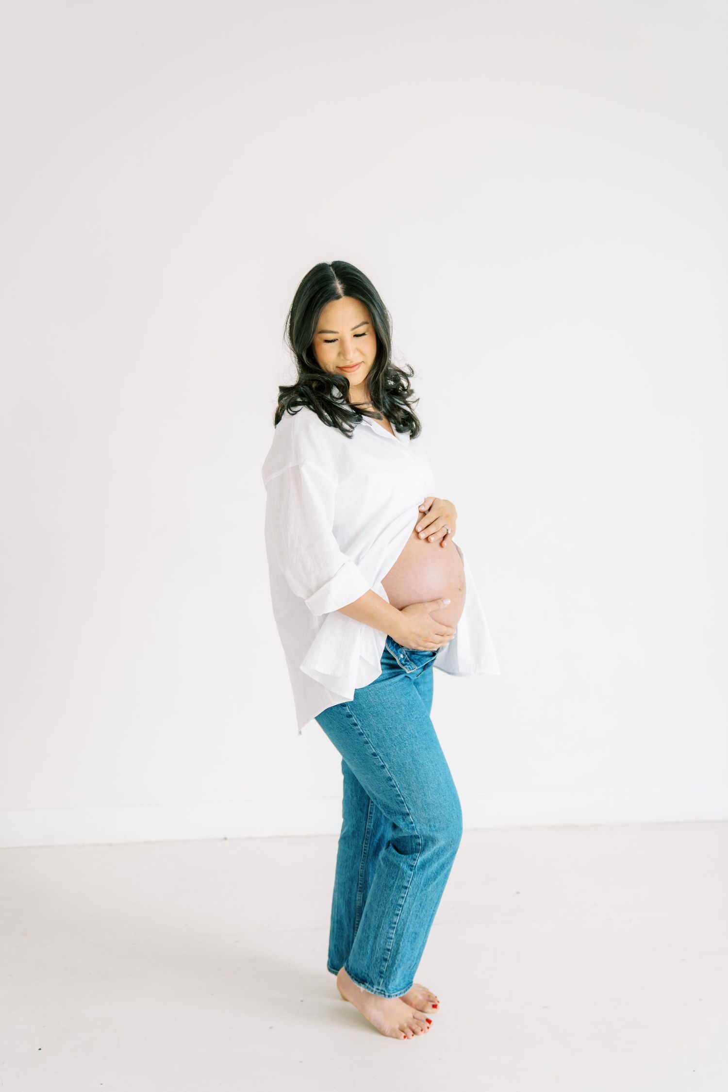 Pregnant woman in white blouse and blue jeans cradling her baby bump against a white background.
