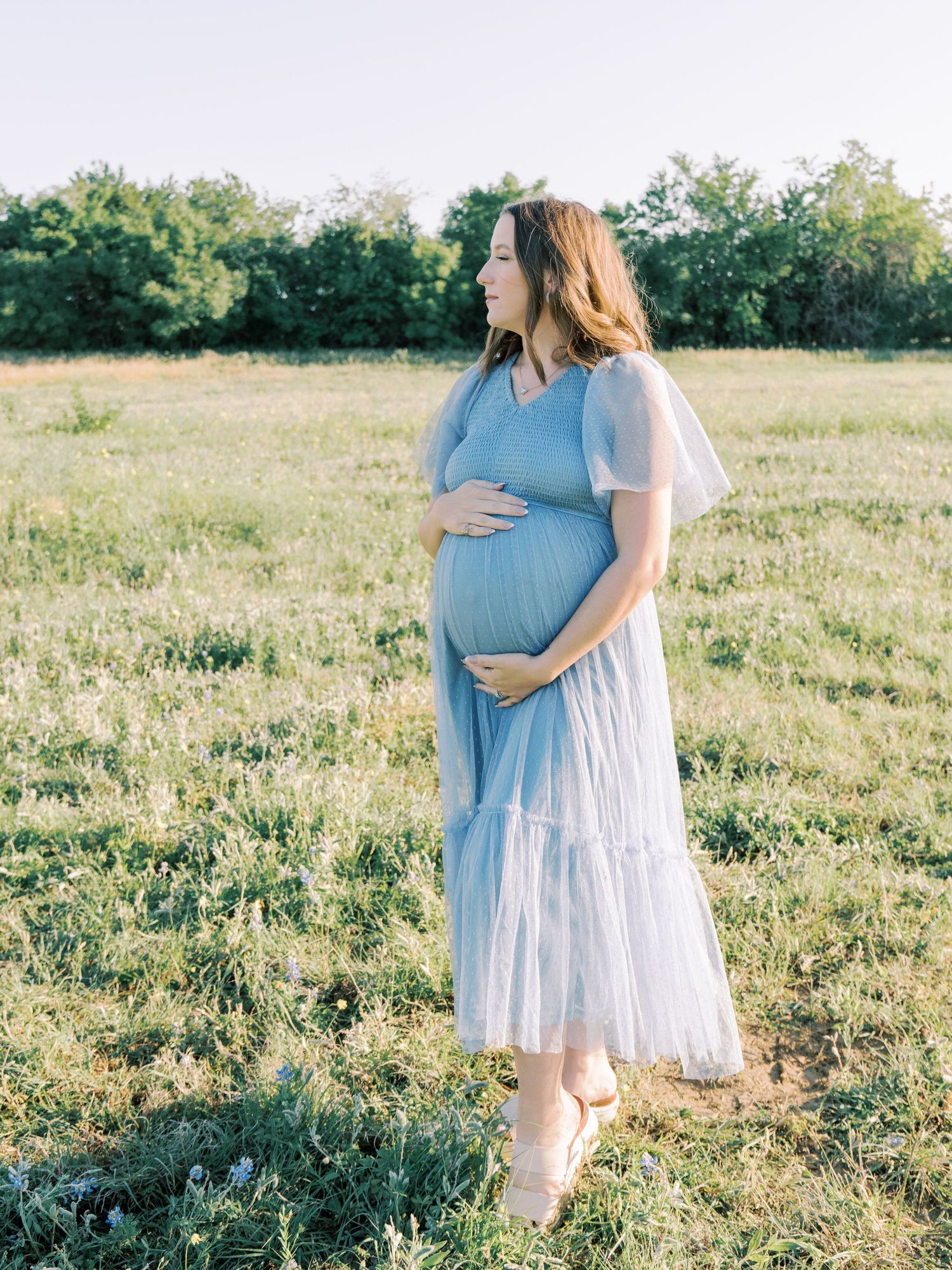 Pregnant woman in light blue tulle dress cradling her baby bump in a sunlit green field.