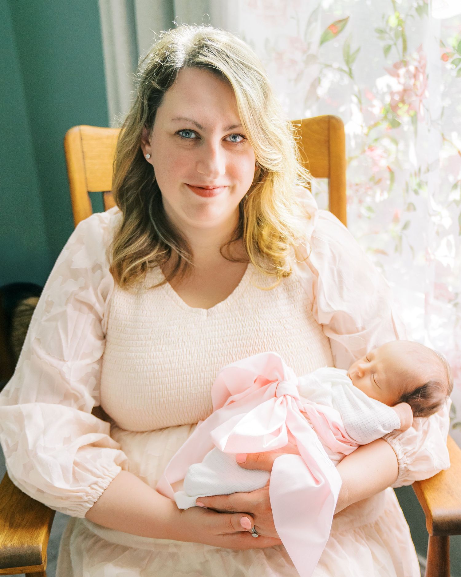 A woman in a pink dress holds a newborn baby wrapped in a pink bow blanket while seated near a window.