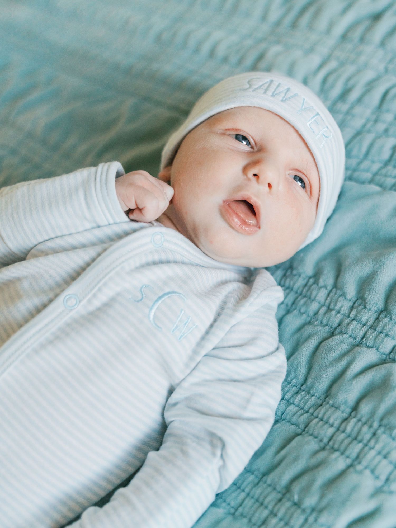 Newborn baby named Sawyer wearing a monogrammed white hat and striped onesie, lying on a teal quilted blanket.