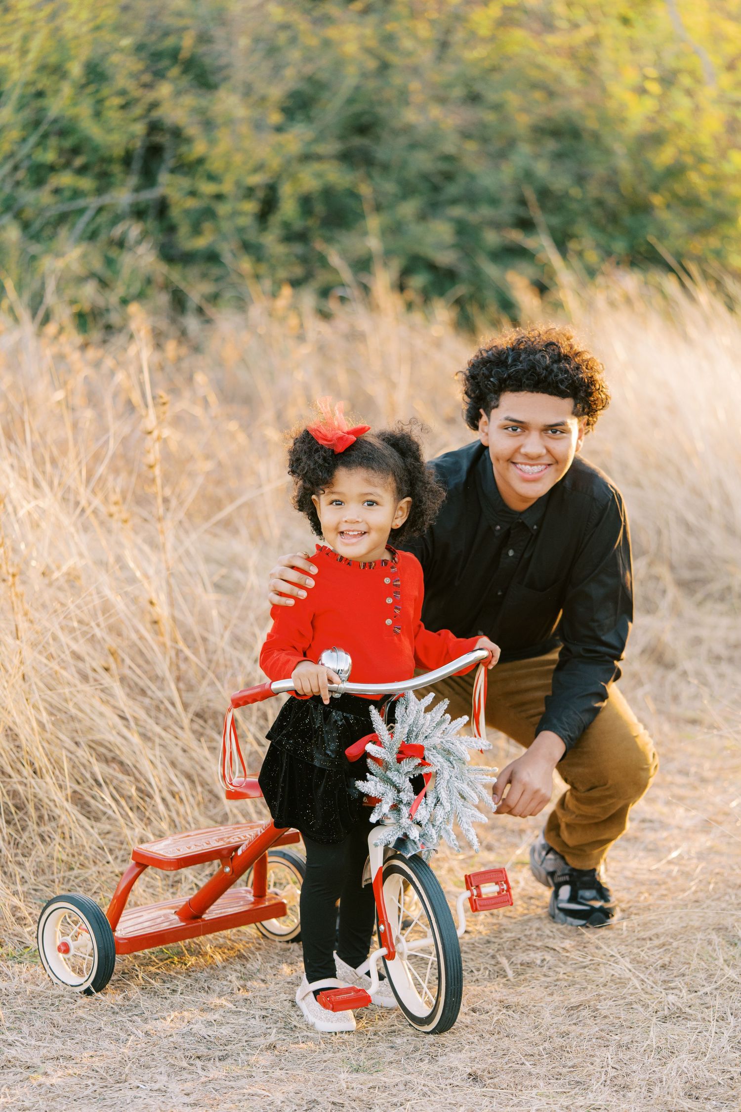 A child rides a small red tricycle in a grassy field while an adult watches and helps.