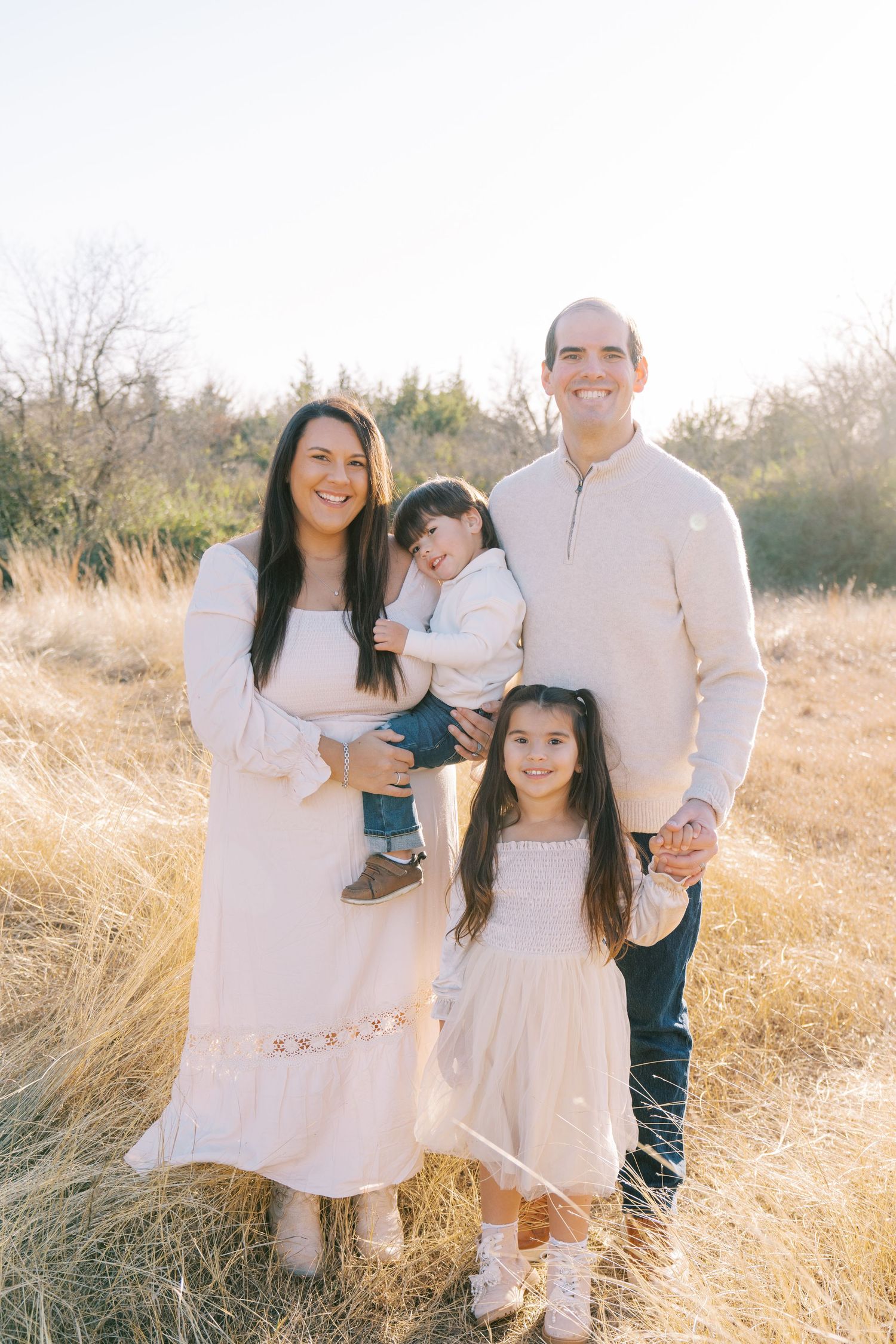 A family wearing coordinated cream-colored outfits poses together in a sunlit field with golden grass.