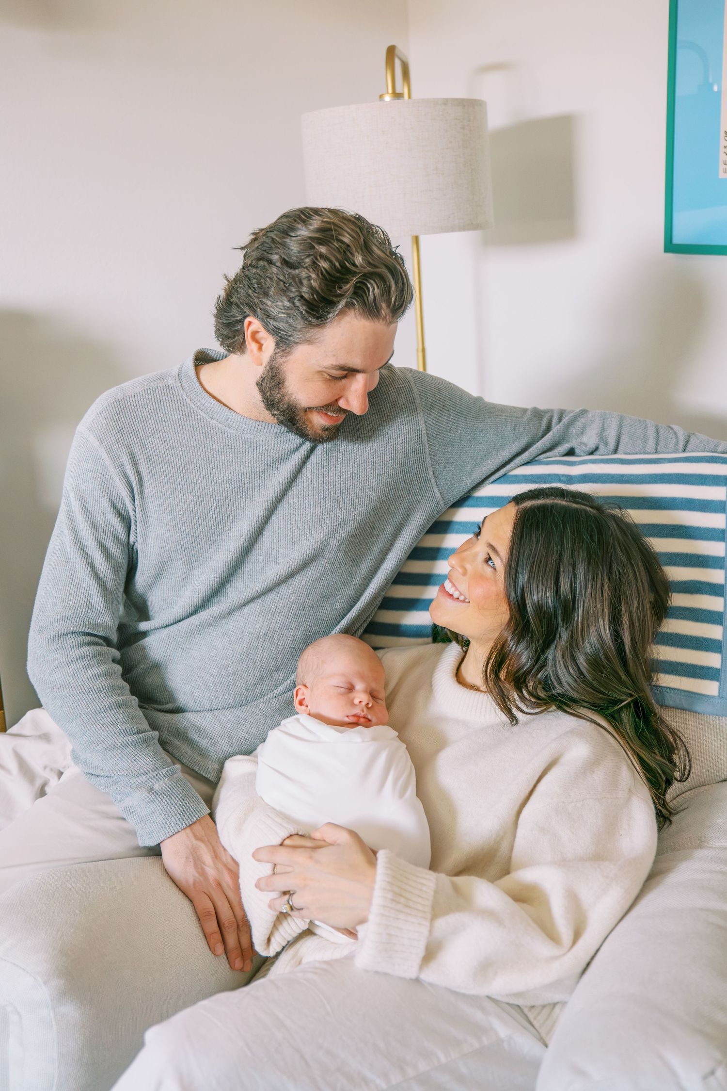 A couple smiles at each other while holding their sleeping newborn baby on a bed with white bedding.