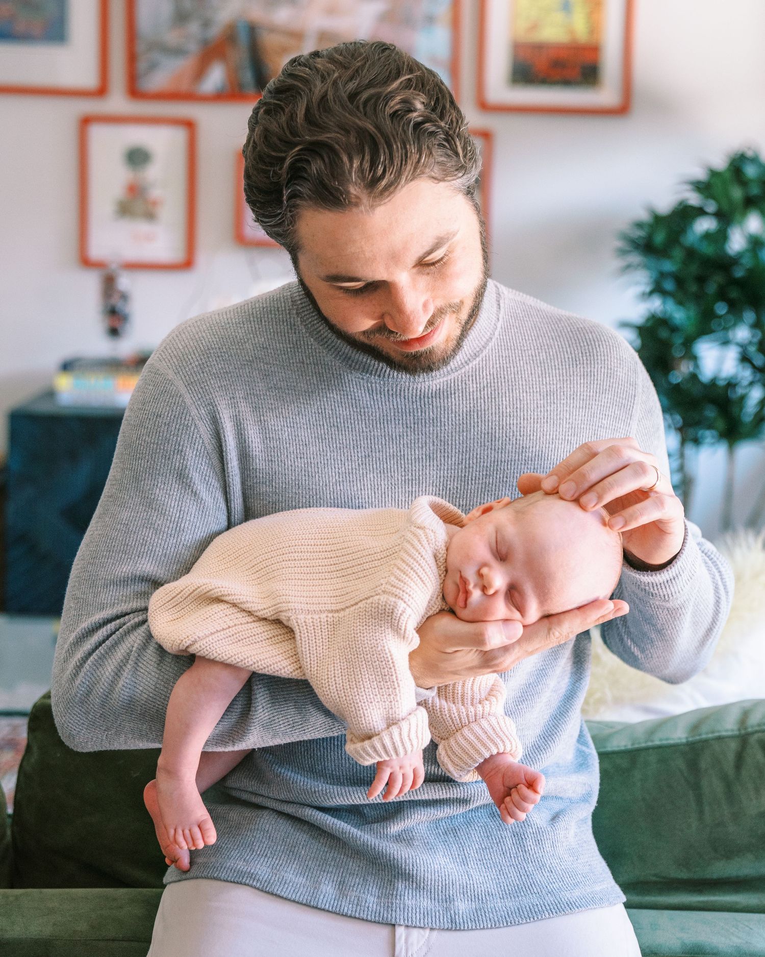 A bearded man in a gray sweater tenderly holds a sleeping newborn baby dressed in a pink knit outfit.