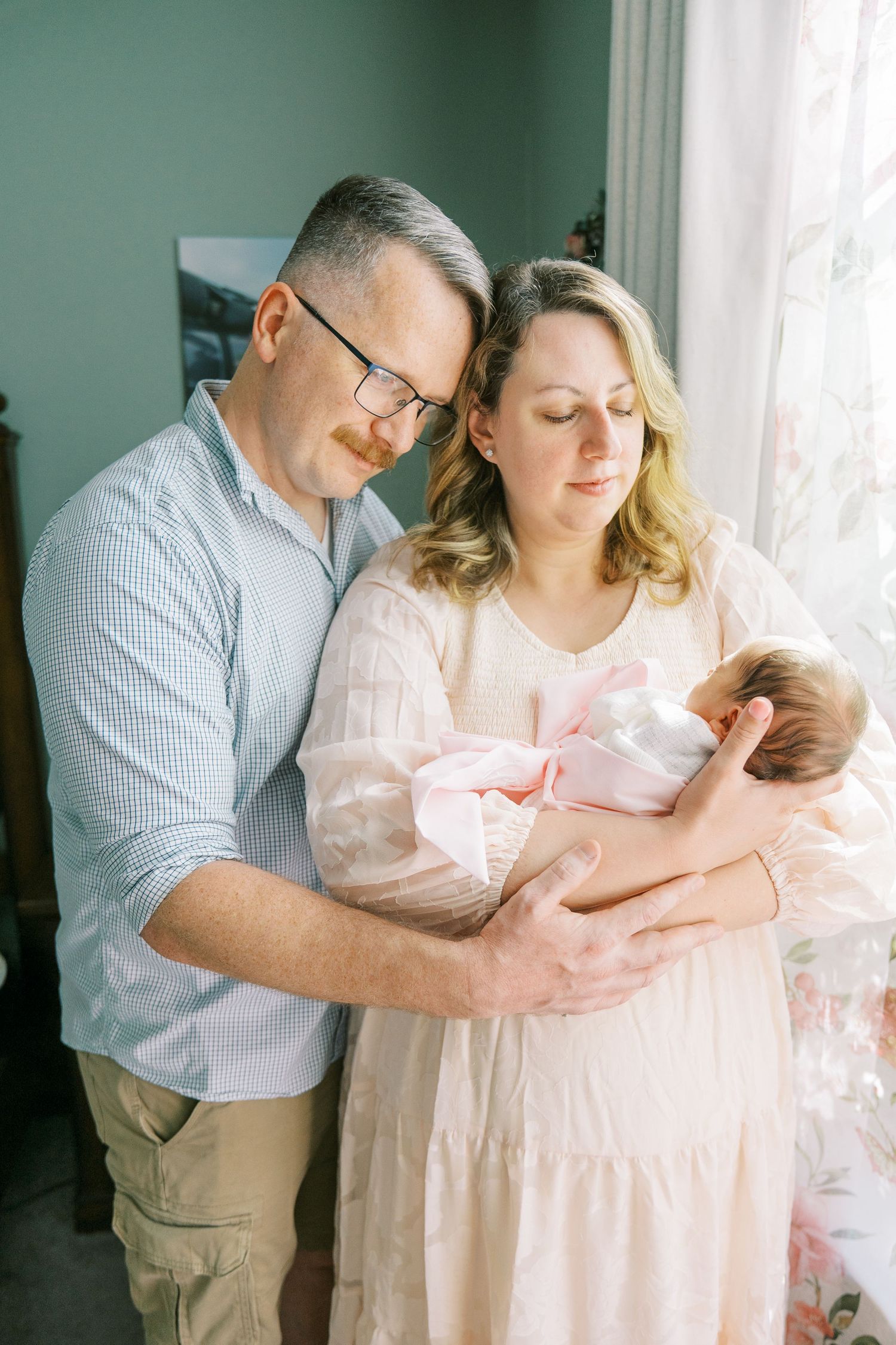Parents in blue and pink outfits lovingly hold their newborn baby near a bright window.