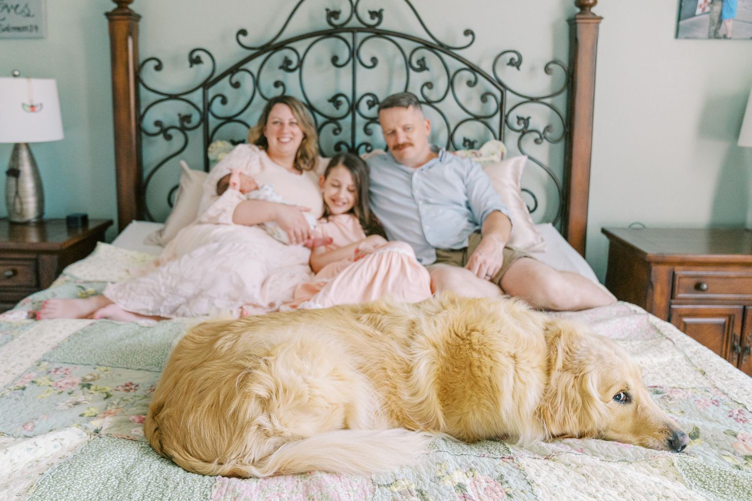 Family lounging on decorative metal bed with golden retriever dog resting at their feet.
