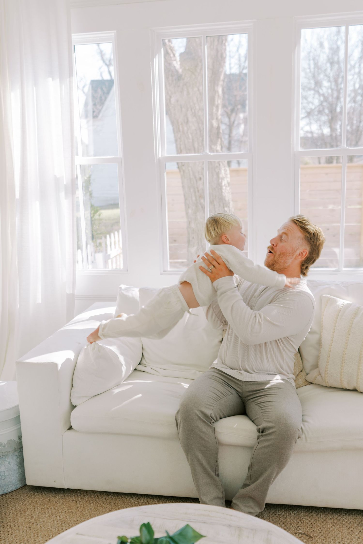 Playful moment on white couch as toddler is lifted up in sunlit room.